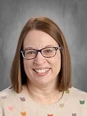 Portrait of a woman with shoulder-length red hair, glasses, and a light-colored sweater with small colorful patterns, smiling against a gray background.