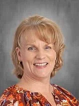 A smiling woman with short blonde hair wearing a colorful patterned blouse, standing against a gray background.