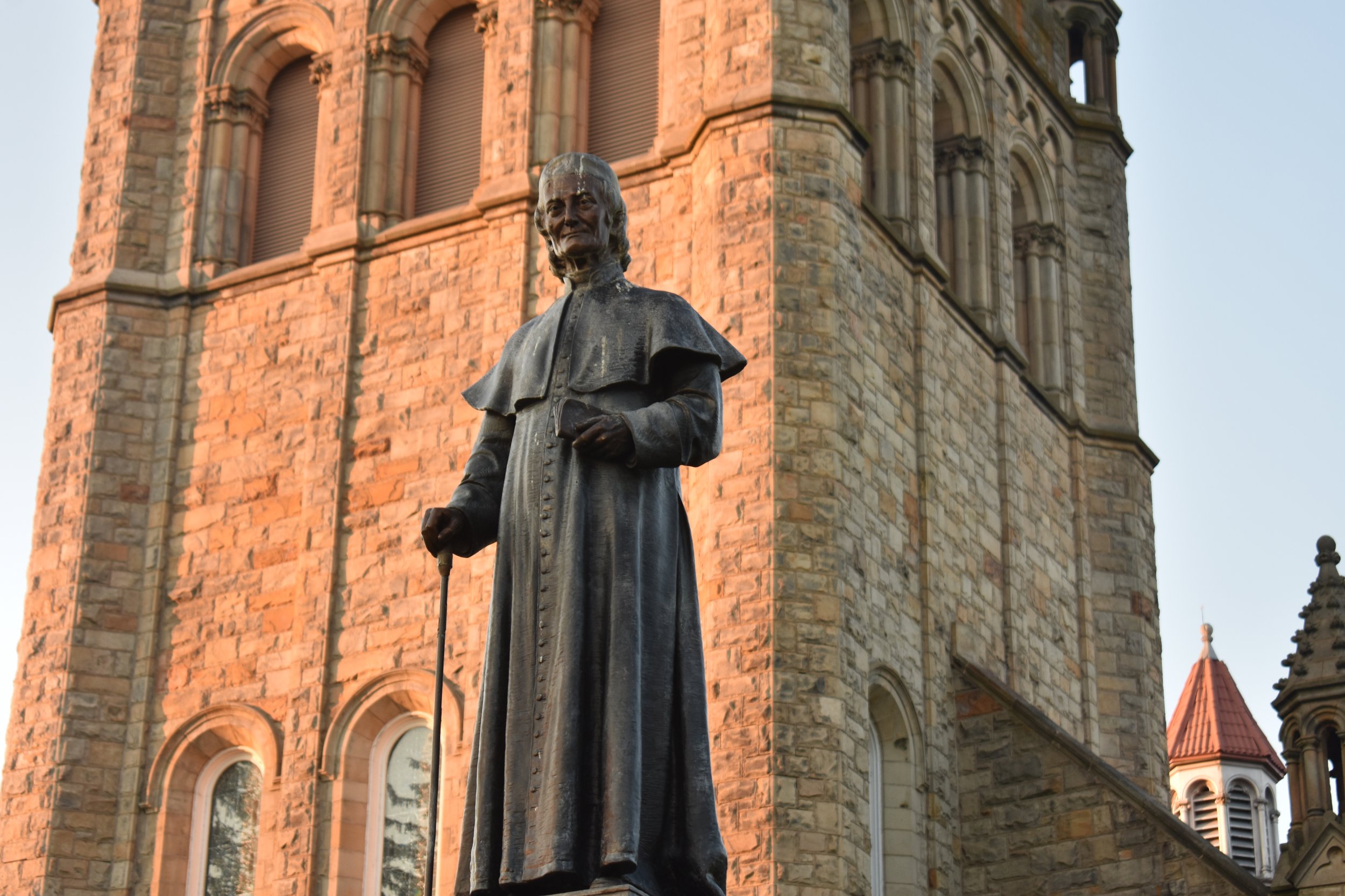 Statue of a historical figure in front of a large, old stone building with arched windows and a tower. The statue depicts a man in period clothing, holding a cane, with a serious expression. The building appears to be made of sandstone or similar material, with detailed architectural features.