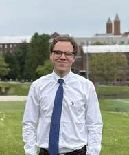 A man in a white shirt and blue tie standing outdoors in front of a grassy area with trees and buildings in the background.