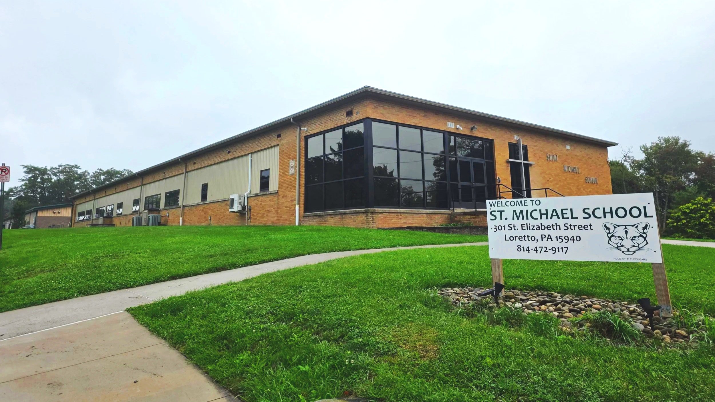The image shows a school building with brick and beige exterior walls, large glass windows, and a at grass lawn in front. There is a white sign with black text and a cougar logo that reads "Welcome to St. Michael School, 301 St. Elizabeth Street, Loretto, PA 15940, 814-472-9117."