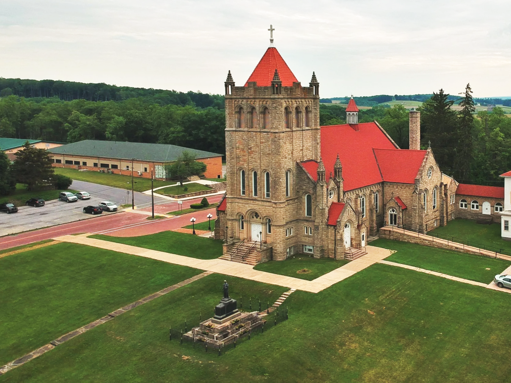 An old stone church with a tall bell tower and red roof, surrounded by a well-maintained lawn, parking lot, and a statue in the foreground, with a forested background and overcast sky.