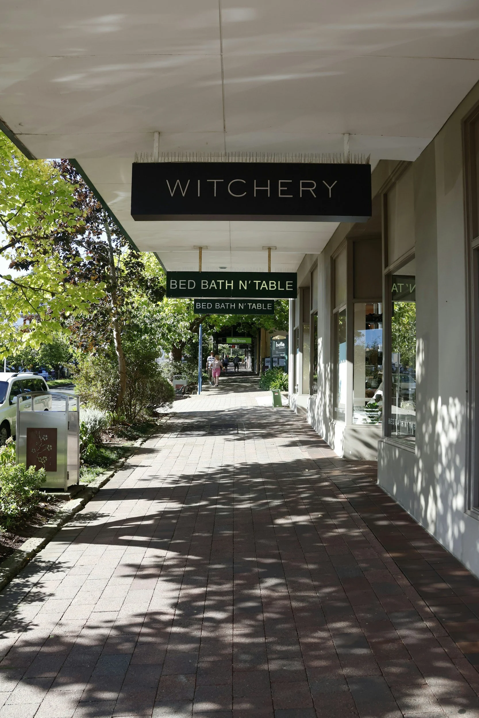 Sidewalk along a storefront with hanging signs reading 'WITCHERY' and 'BED BATH N' TABLE,' greenery, trees, and pedestrians in the background.