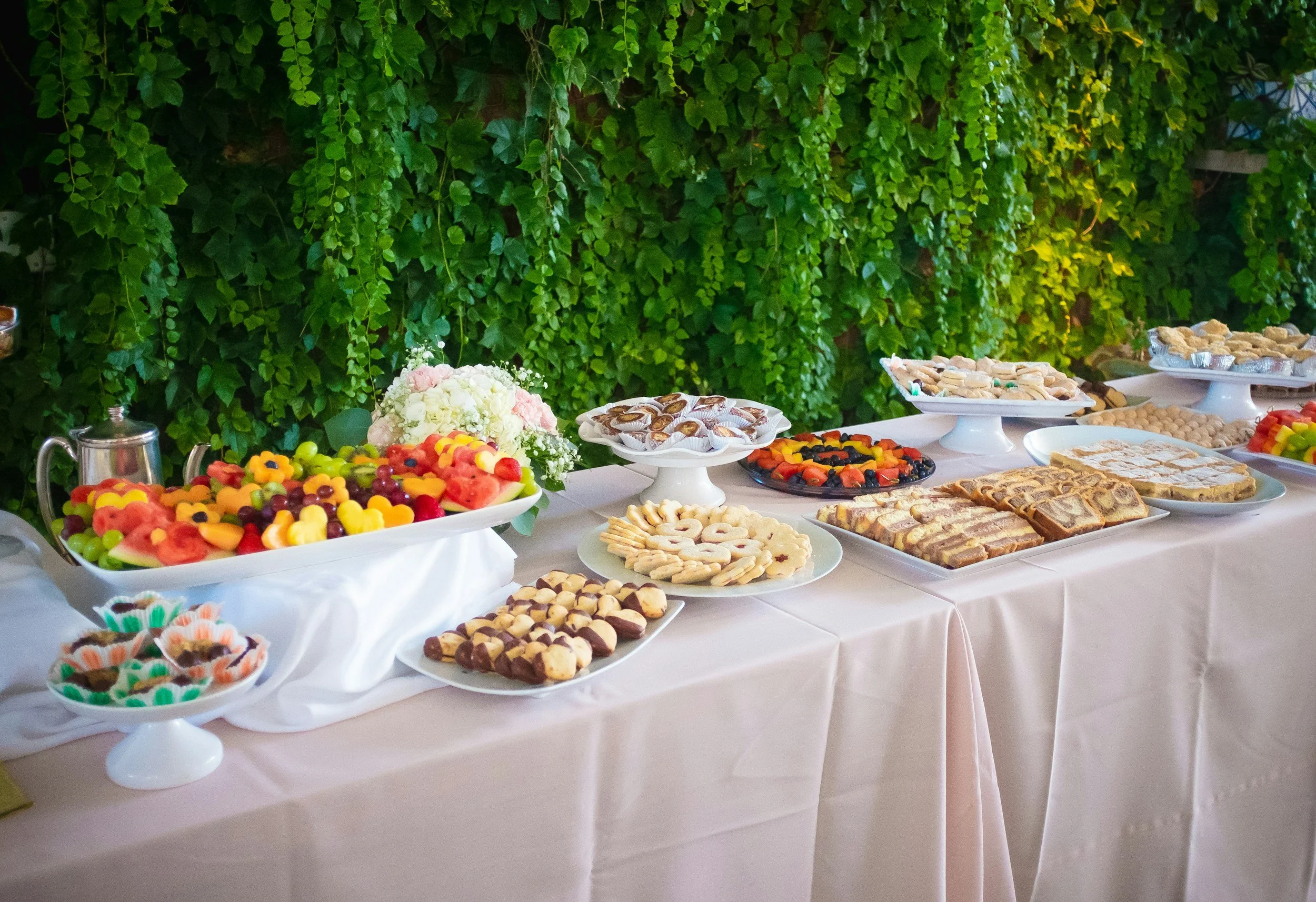 A dessert table filled with various cookies, candies, fruit slices, and a flower arrangement, set against a green leafy background.