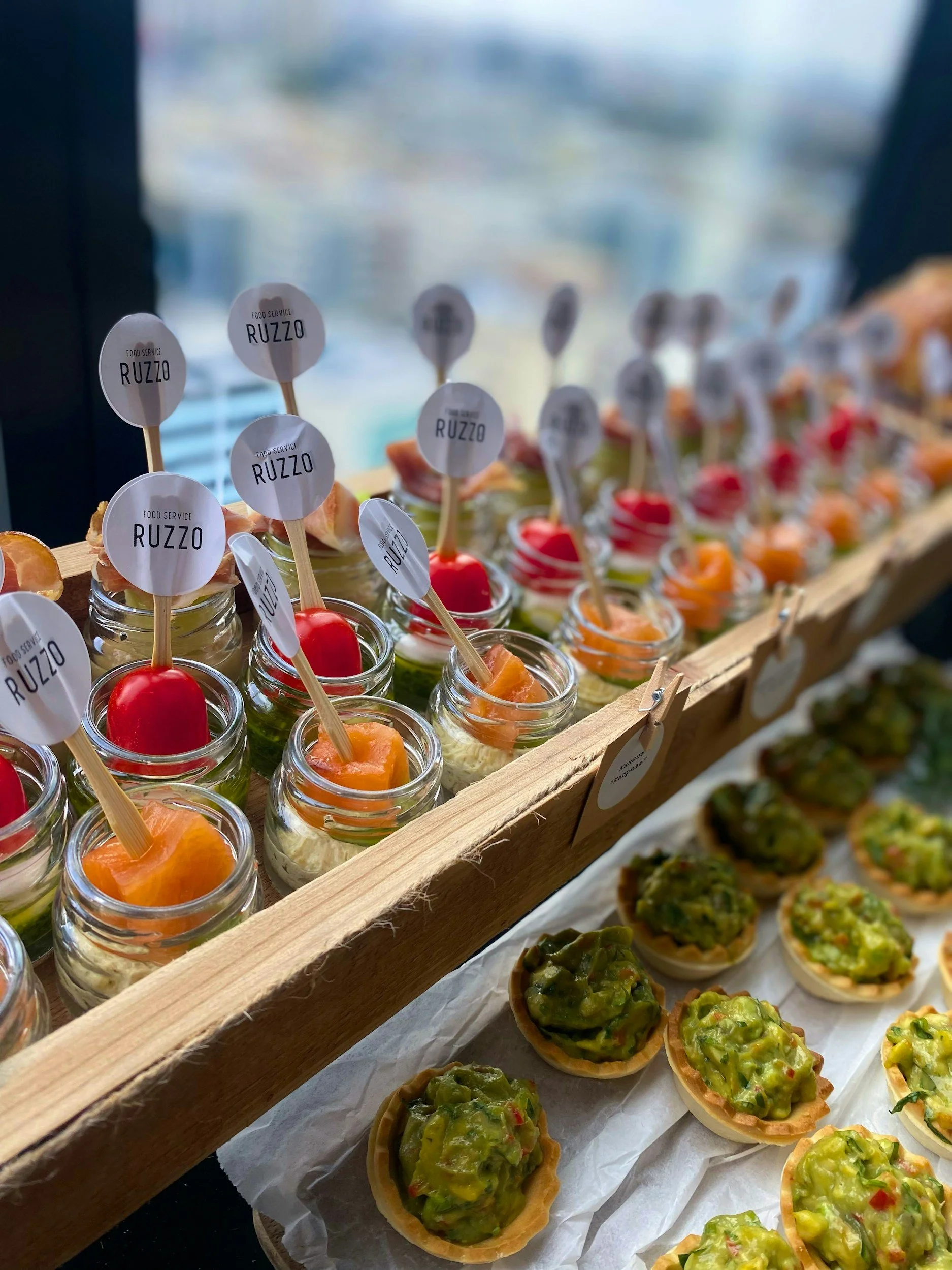 A display of appetizers including small jars with toppings and topped tart shells with green and red vegetables, at a food event or market.
