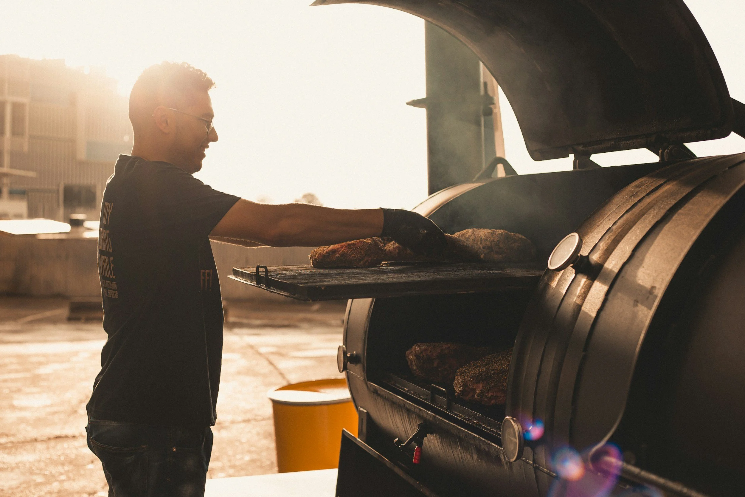 A man wearing glasses and a black t-shirt is standing beside a large industrial oven, placing food inside. The setting is outdoors with sunlight creating a silhouette effect, and a yellow trash can and some buildings are visible in the background.