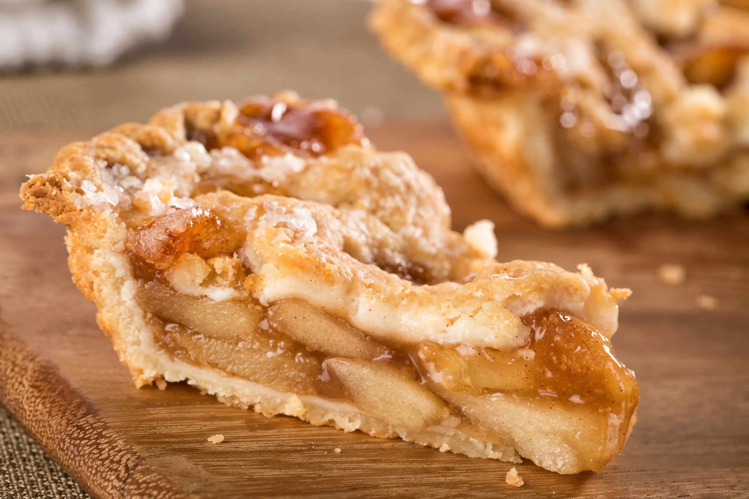 A slice of apple pie with caramel, ice cream, and apple slices on top, placed on a wooden serving board.