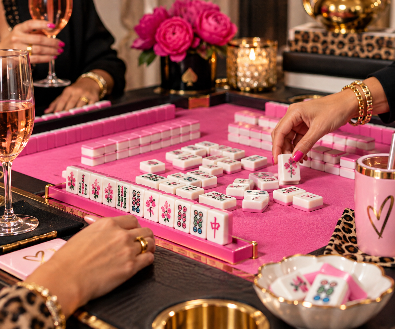 People playing Mahjong at a decorated table with pink and gold accents, pink flowers, and glasses of rose wine.