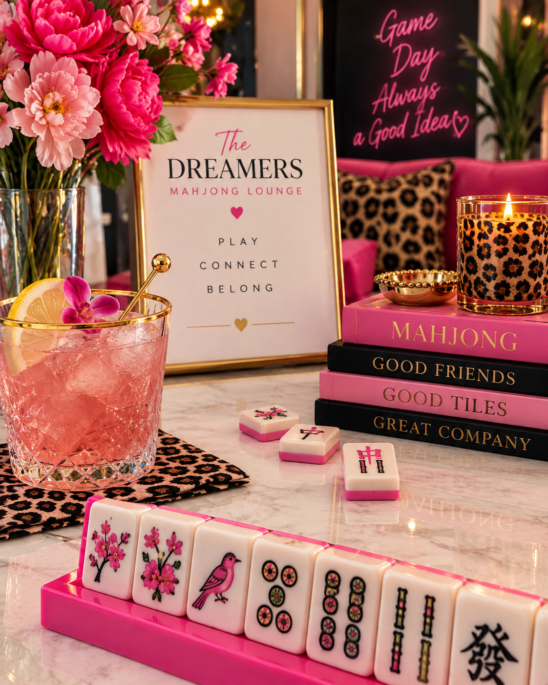 Decorative mahjong lounge setup with pink and black color themes, featuring a vase of pink flowers, a pink cocktail with lemon and orchid garnish, stacks of mahjong and color-themed books, a leopard print candle, and mahjong tiles on a pink tray.