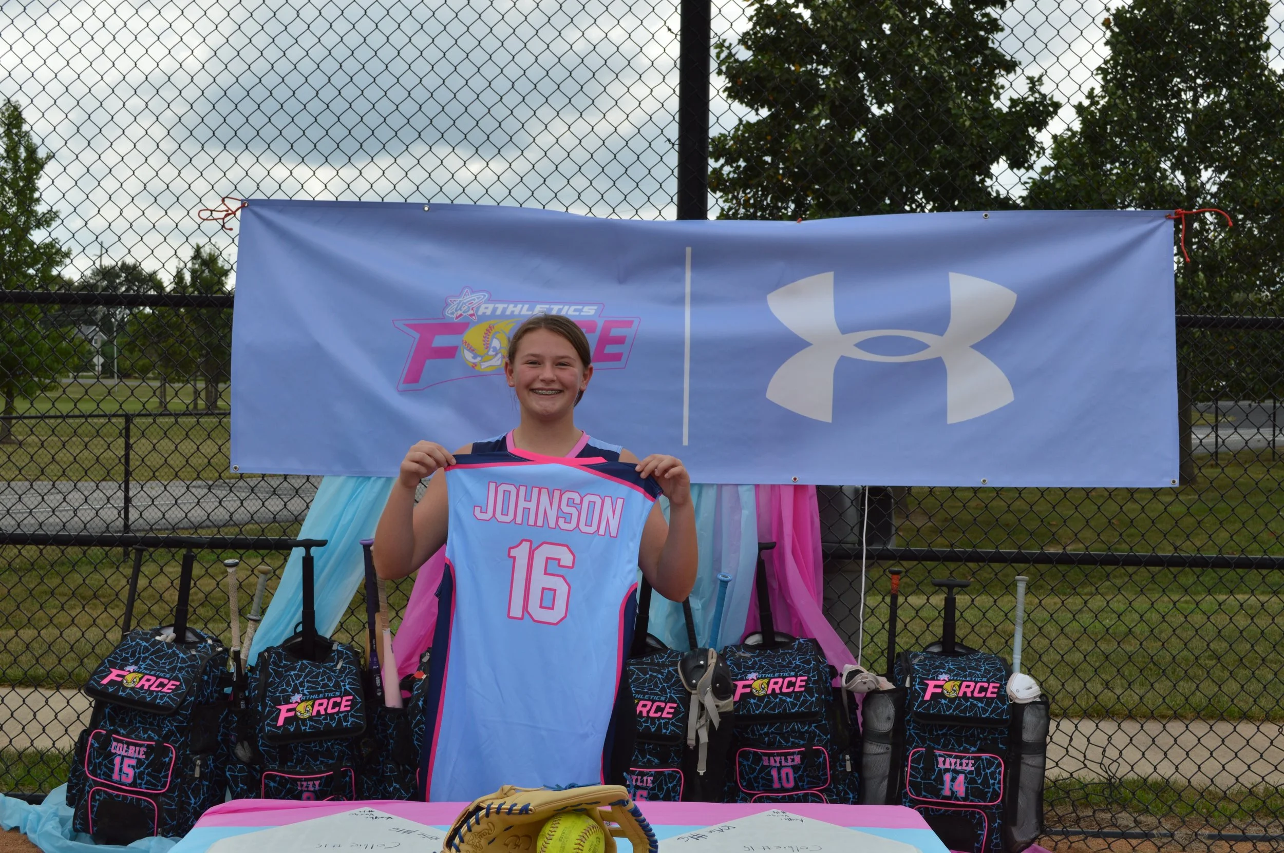 A young girl smiling while holding a volleyball jersey that says Johnson with the number 16. She is standing in front of a table with sports equipment and bags, with a backdrop of a fence, trees, and cloudy sky.
