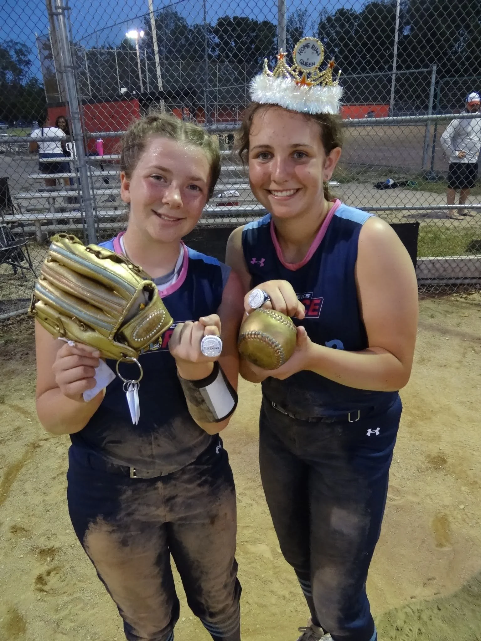Two young women in baseball uniforms celebrating on a baseball field after a game. They are holding a gold baseball and a gold glove, smiling with medals on their necks. One is wearing a decorated 'Home Run Queen' crown. The background includes a chain-link fence and others in the dugout area.