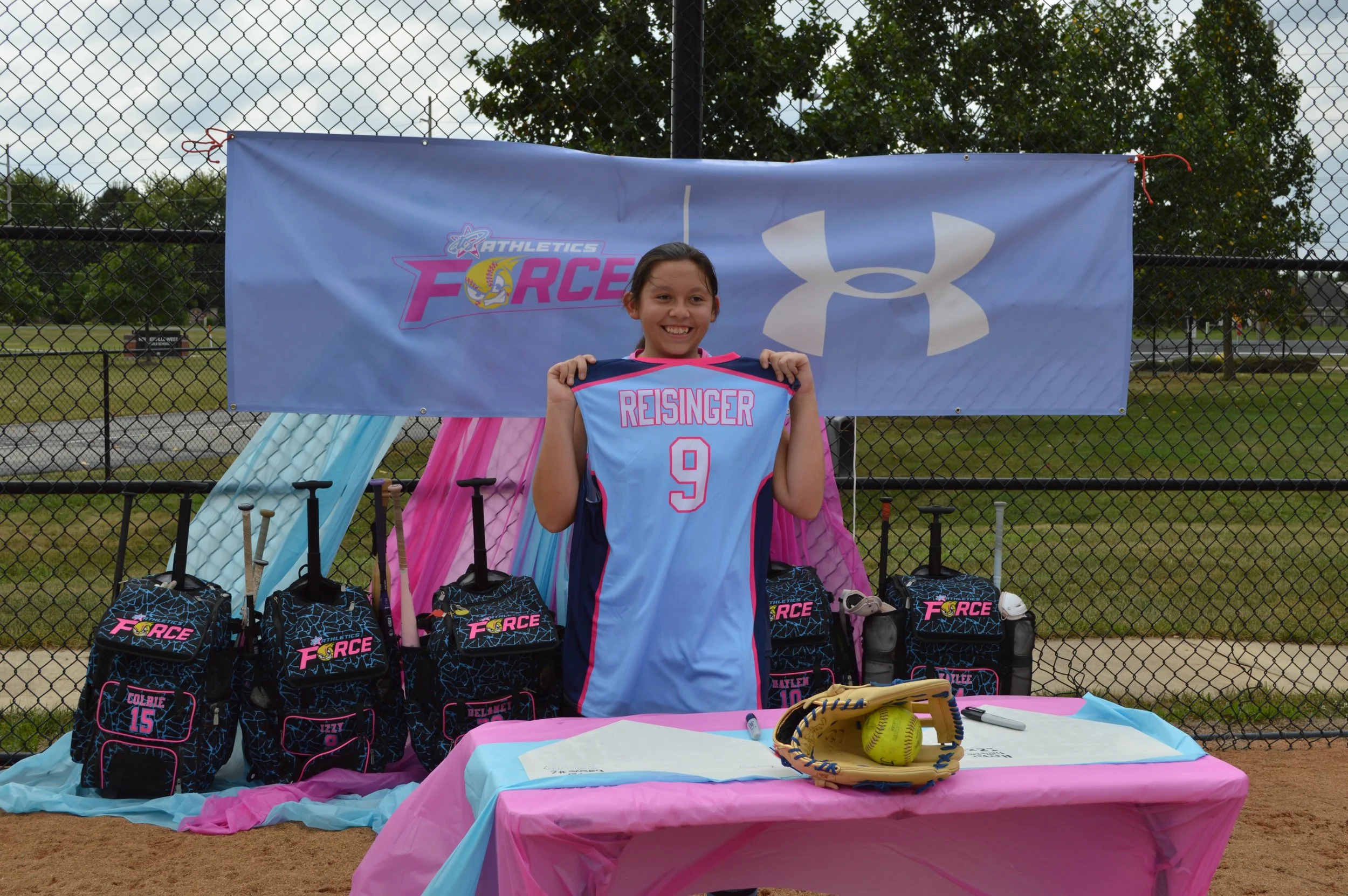 A young girl holding a light blue sports jersey with the name 'RESINGER' and the number 9, standing in front of a table and backdrop at a sports event. The table has pink and blue tablecloths, with baseball gloves, a ball, and markers. The backdrop features a large logo for 'Athletics Force' and a prominent Under Armour logo. There is a fence and trees in the background.