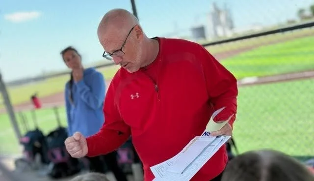A man in a red jacket with glasses holding papers, celebrating or feeling triumphant, at a sports field or track with a woman in the background
