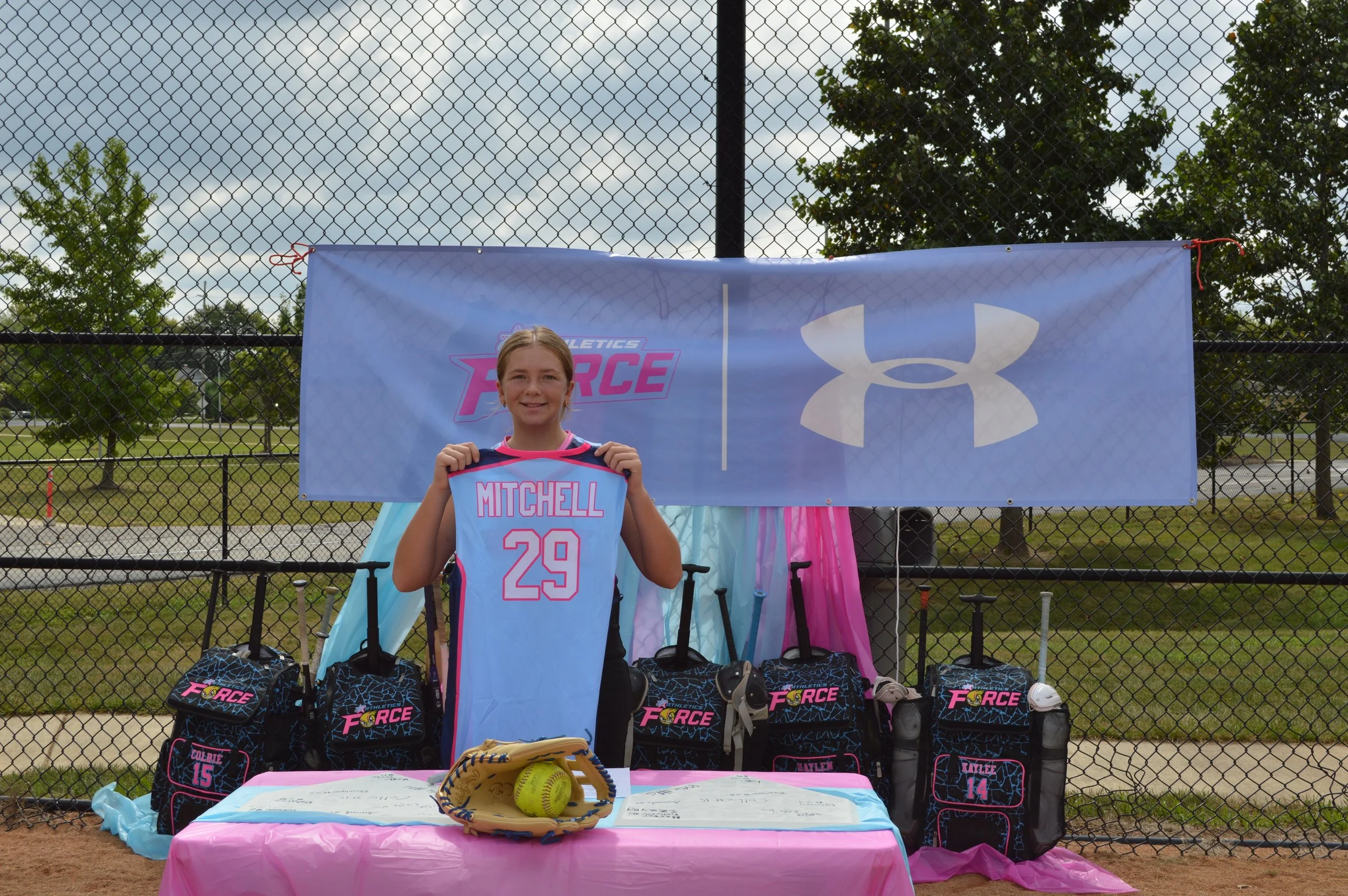 A young girl standing in front of a sports booth at a baseball field, holding a pink and blue sports jersey with the number 29 and the name Mitchell. The booth displays several black backpacks with pink and purple lettering reading "Force", along with baseball gloves and balls. The backdrop includes a blue banner with a white logo and the text "Athletics Force", behind a chain-link fence during daytime.