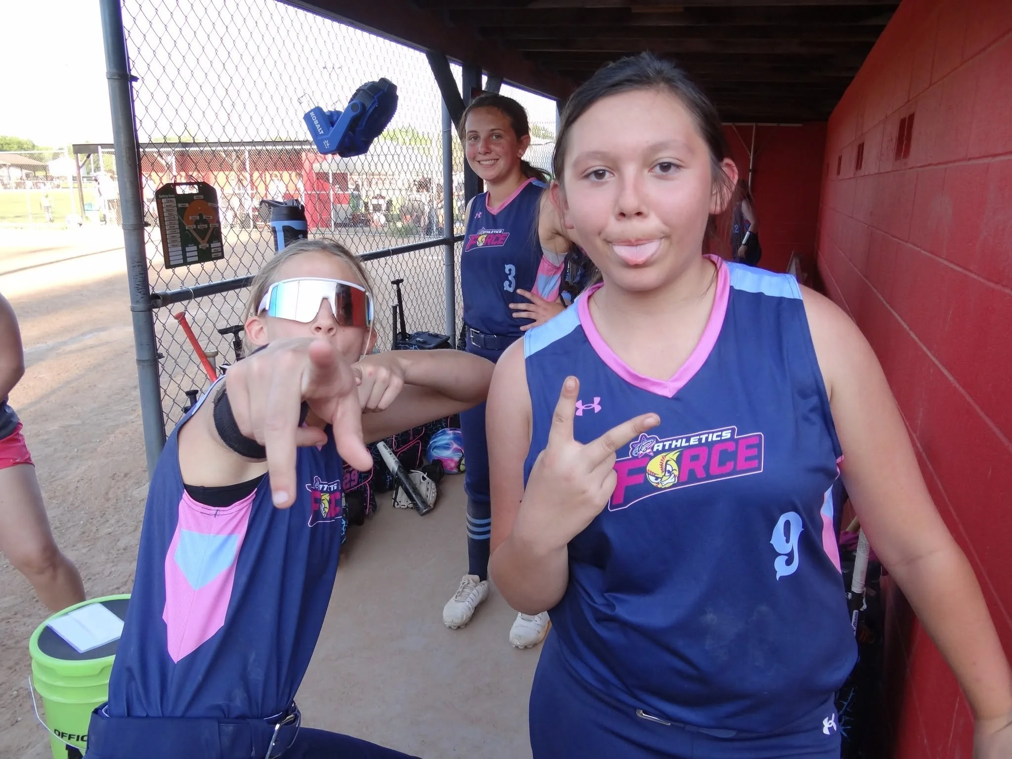 Three young girls in sports uniforms at a baseball field, with two of them making playful gestures toward the camera. One girl is sticking out her tongue, and another is pointing her finger. The background shows a chain-link fence, a red dugout wall, and spectators and players outside the field.