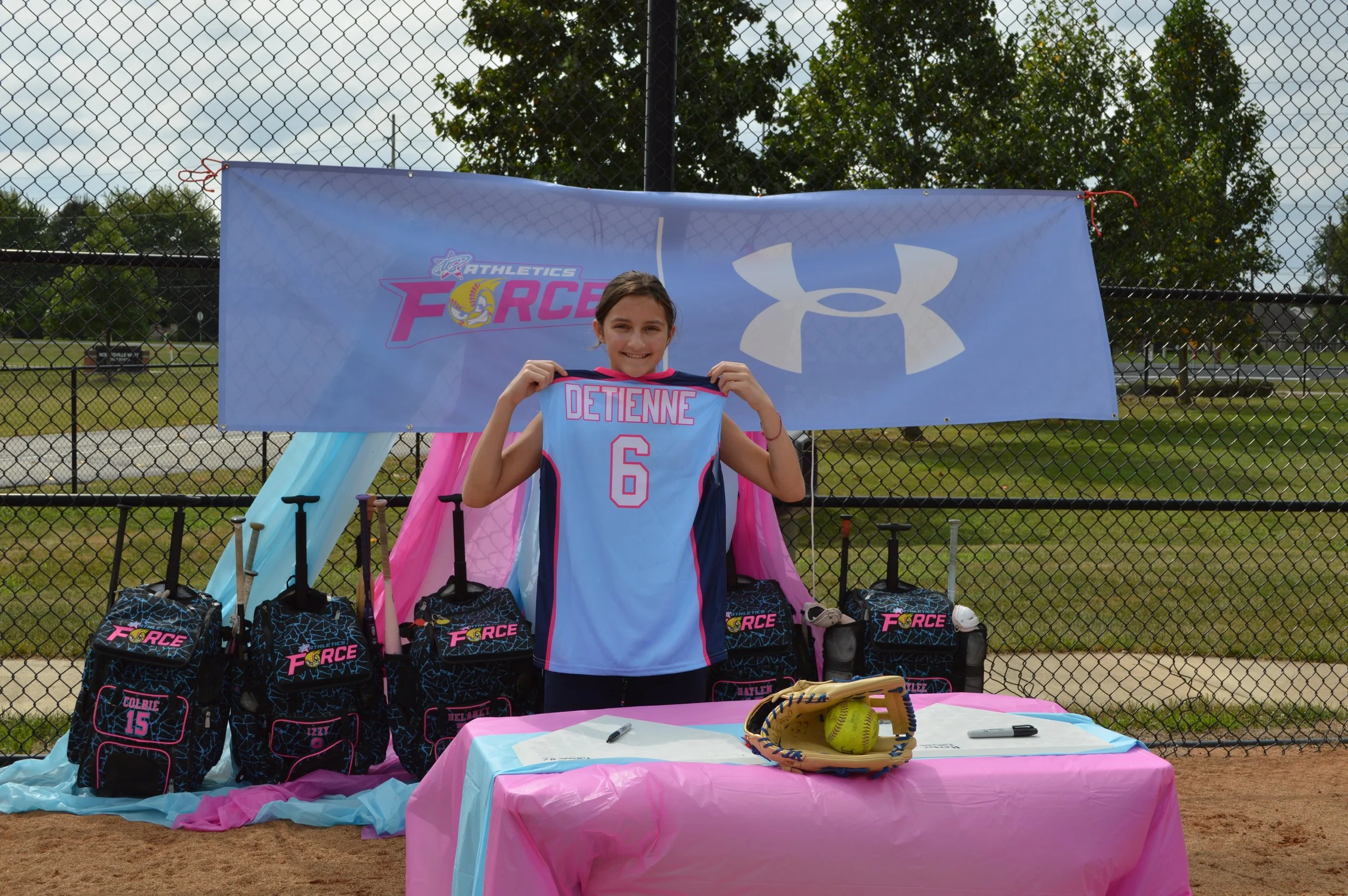 A young girl in a sports uniform holds a jersey with the name DETIENNE and the number 6. She is smiling and standing in front of a table with sports equipment and a backdrop with sports logos, on a fence at a sports field.