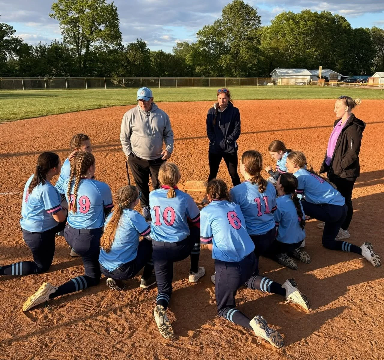 A girls' softball team huddles on the field during a game with their coaches standing nearby. The team is wearing matching light blue uniforms with numbers on the back and kneeling on the dirt infield.