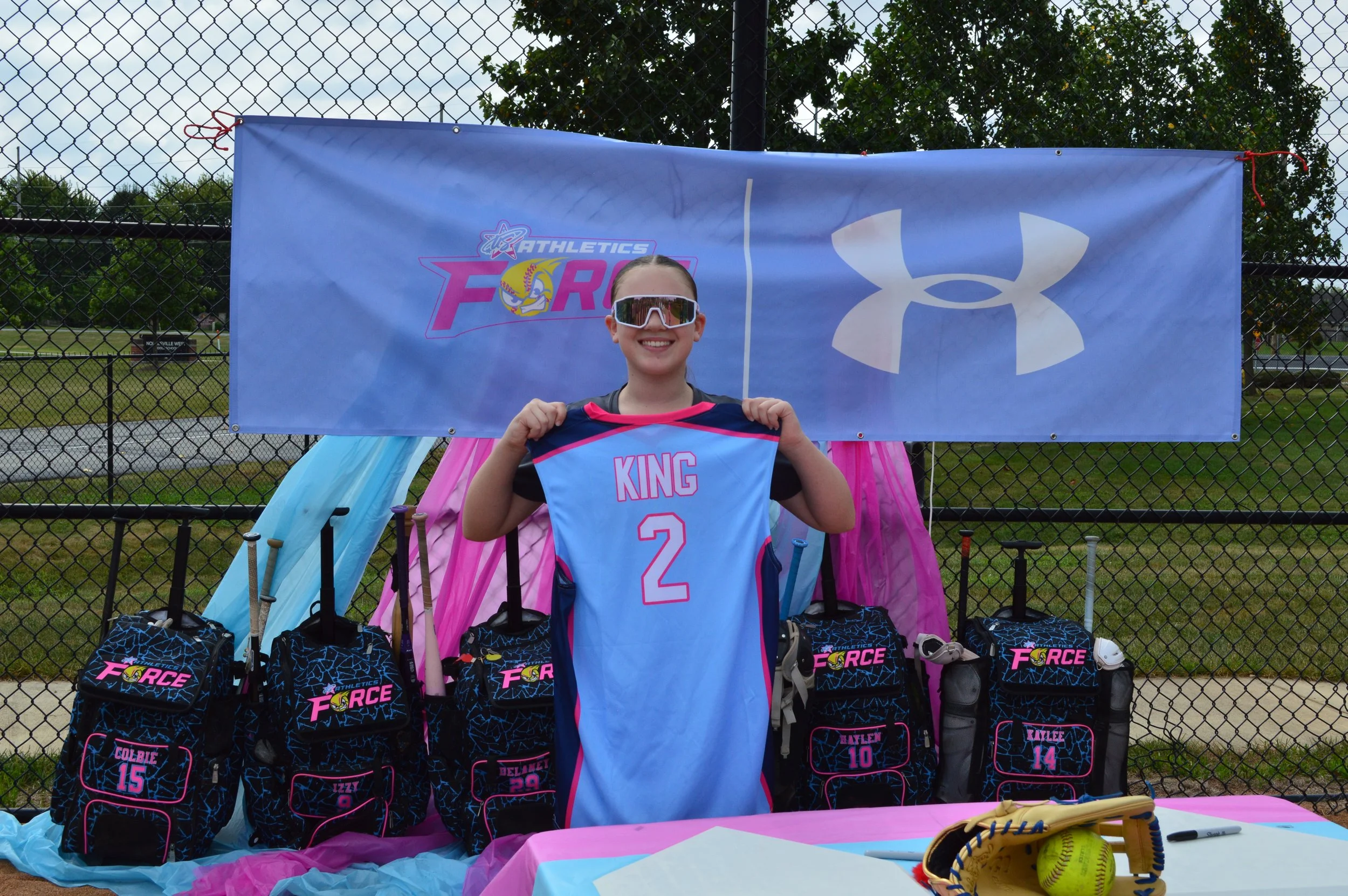 A young girl with sunglasses smiling and holding a sports jersey that says 'KING' and the number 2. She is standing behind a display of softball gear and bags, with a blue banner behind her that features the logos of Athletico Force and Under Armour. The scene appears to be at an outdoor sports event.