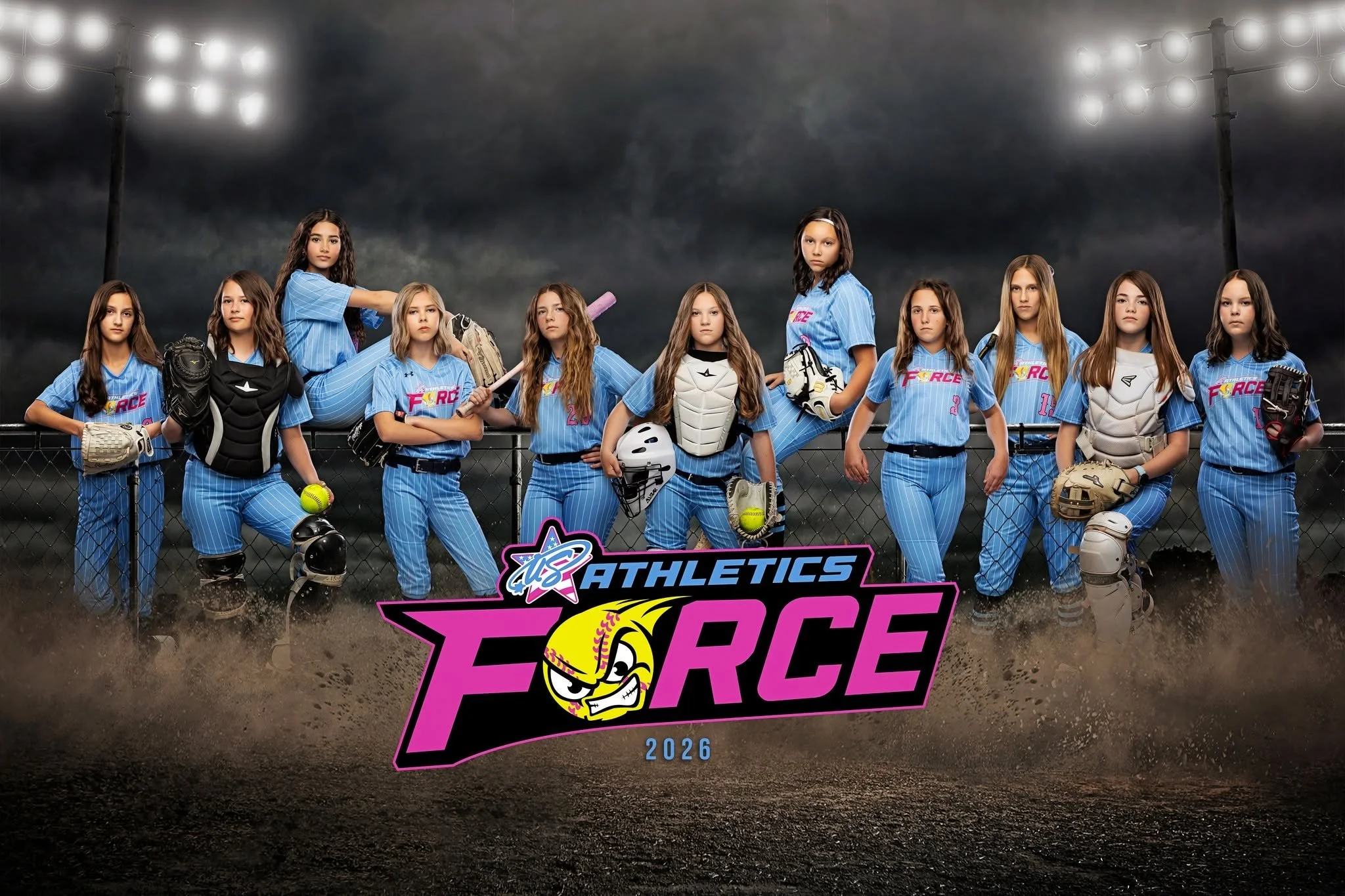 A group of young girls in blue baseball uniforms standing on a baseball field with a dark, cloudy sky in the background. They are posing behind a fence with a banner that reads 'USA Athletics' and 'Force 2026'.