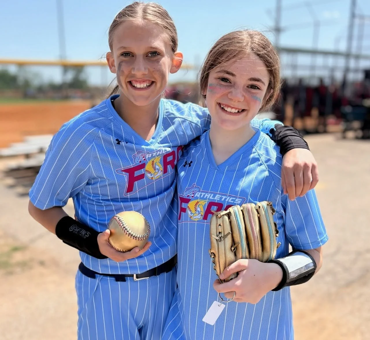 Two young female softball players in blue striped uniforms smiling and posing together on the field. One is holding a softball, and the other has a baseball glove.
