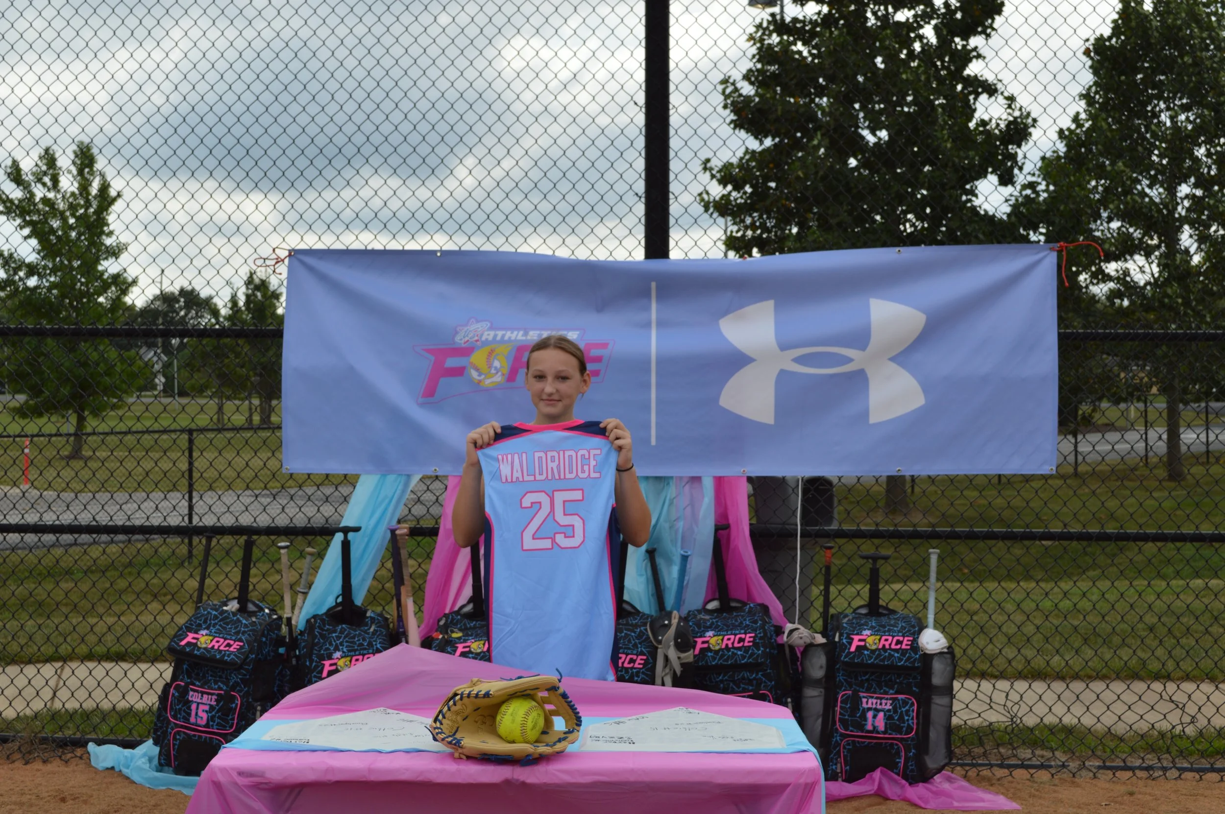 Young girl standing in front of a table at a sports event, holding a pink and blue sports jersey with the name 'WALDRIGE' and the number 25. Behind her are sports bags, a yellow softball in a glove on the table, and a backdrop with logos, all set on a fenced sports field with trees in the background.