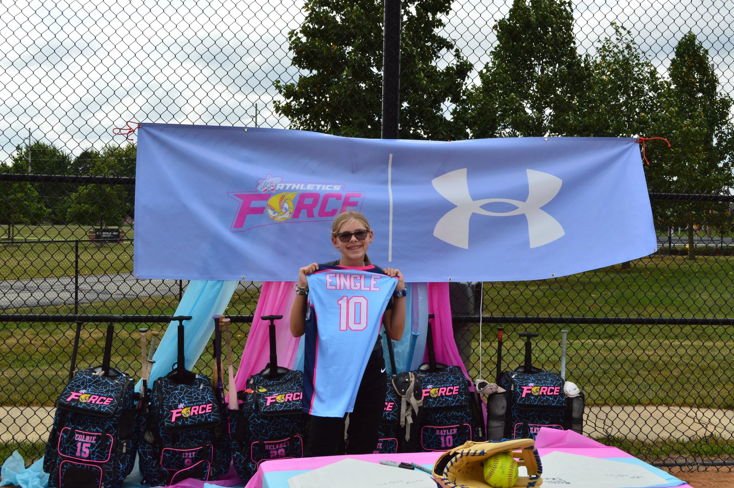 Youth girl standing in front of a colorful athletics themed display outside on a cloudy day, holding a light blue sports jersey with the name 'EINGLE' and number '10'. The display features a large Under Armour logo, an 'Athletics Force' banner, and backpacks with the same branding. Several baseballs and sports equipment are on a table in front.