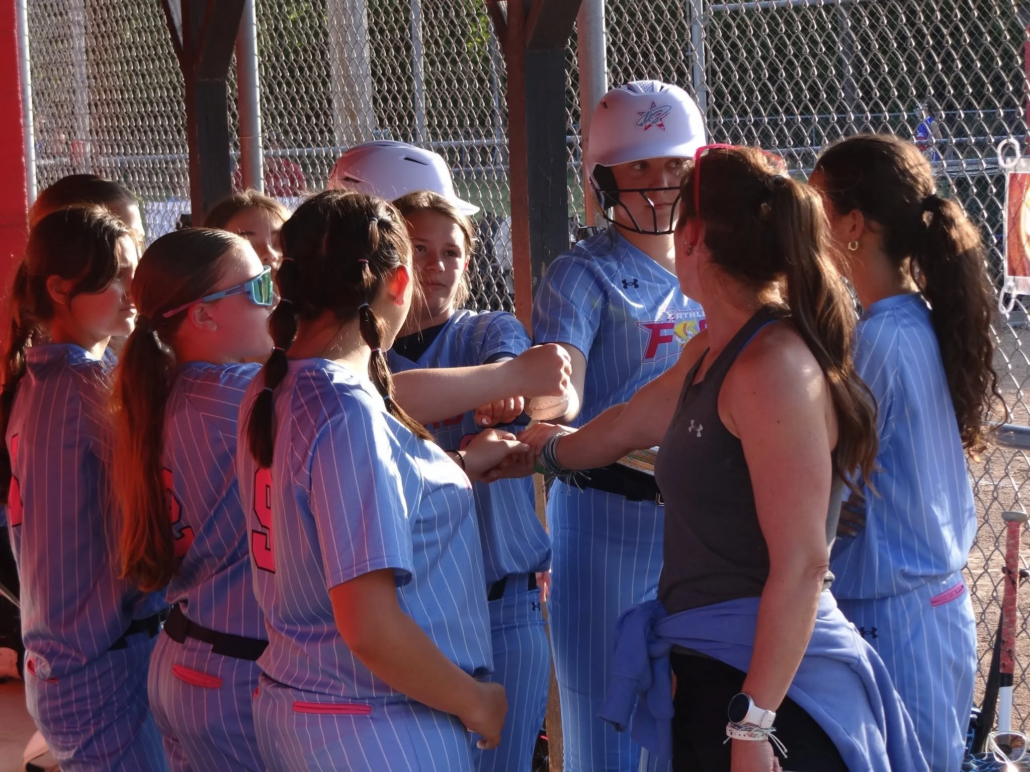 A group of young girls in baseball uniforms and a woman, possibly a coach, are having a moment of celebration at a baseball field. The girls are standing close together, some wearing protective eye goggles, and the woman is hugging one of the players. They are near a chain-link fence, with a batter's box and batting cage visible in the background.
