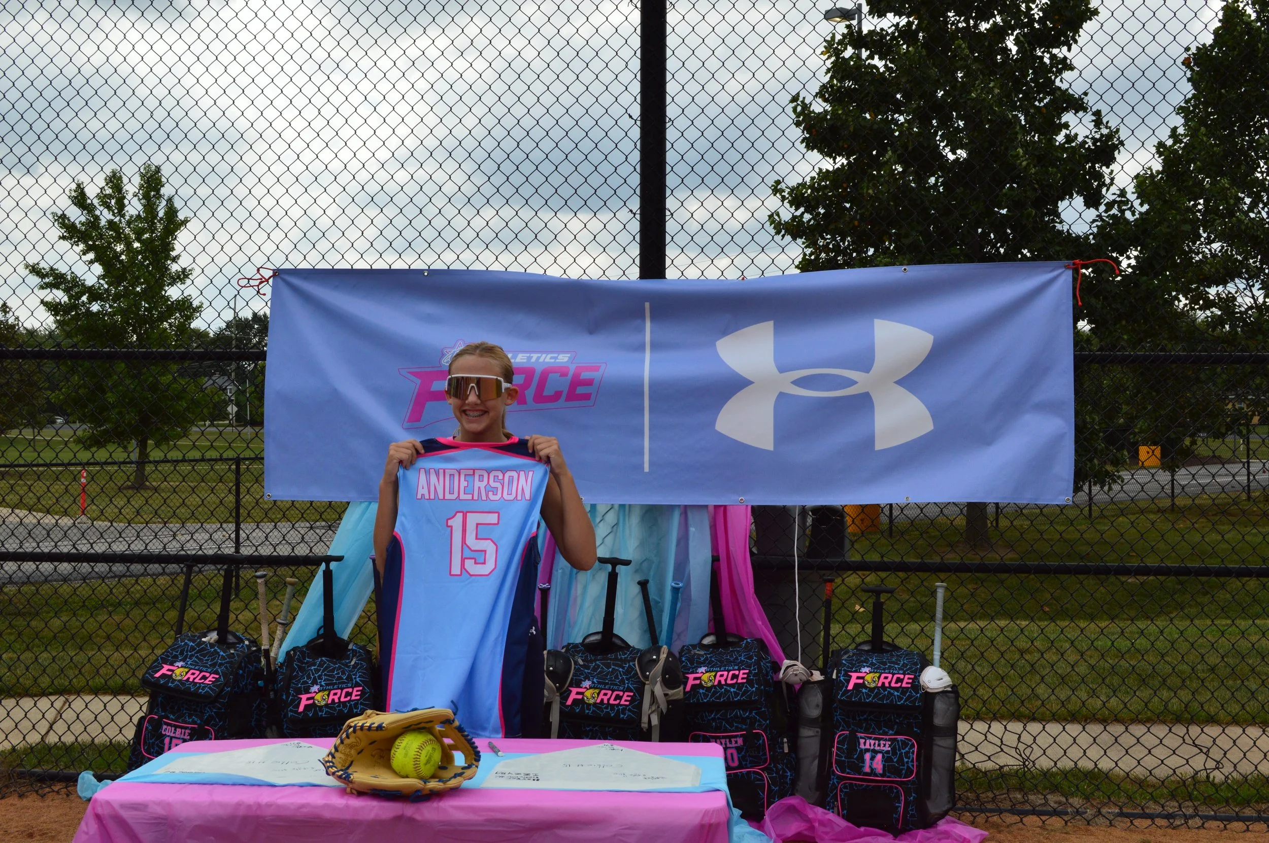 A young girl standing behind a table at a softball event, holding up a blue softball jersey with the name 'Anderson' and the number 15. She is wearing sunglasses and smiling. The table has softball equipment and promotional materials. Behind her is a fence with a banner that has the Under Armour logo and the words 'Athletics Force,' and several sports bags labeled 'Force.'