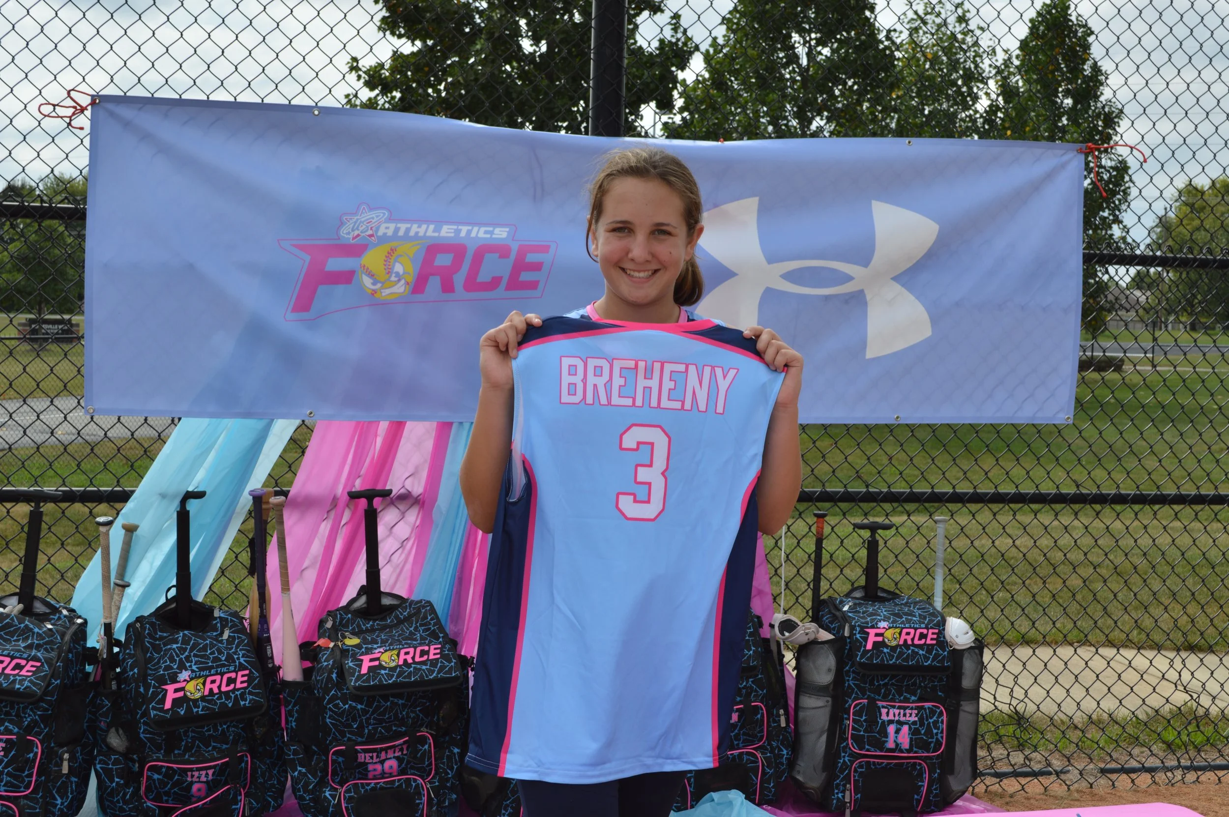 Young girl standing behind a sports gear display at a sports event, holding a blue jersey with pink lettering. Behind her is a banner with the logos of Athletics Force and Under Armour, a chain-link fence, and trees in the background.