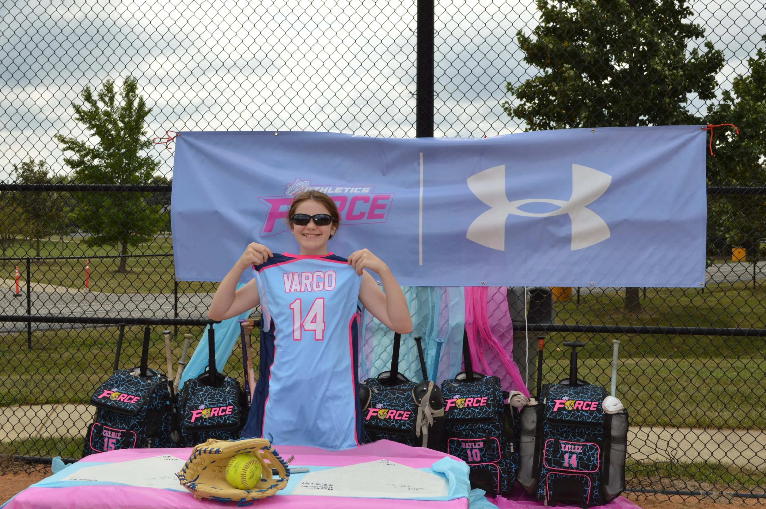 A young girl with sunglasses smiling, holding a blue sports jersey with the name Vargas and the number 14. Behind her, there are sports bags with the 'Athletics Force' logo displayed on a fence at a sports field. The table in front has a yellow softball and a glove, with pink and blue wraps or fabric. Trees and a cloudy sky are visible in the background.