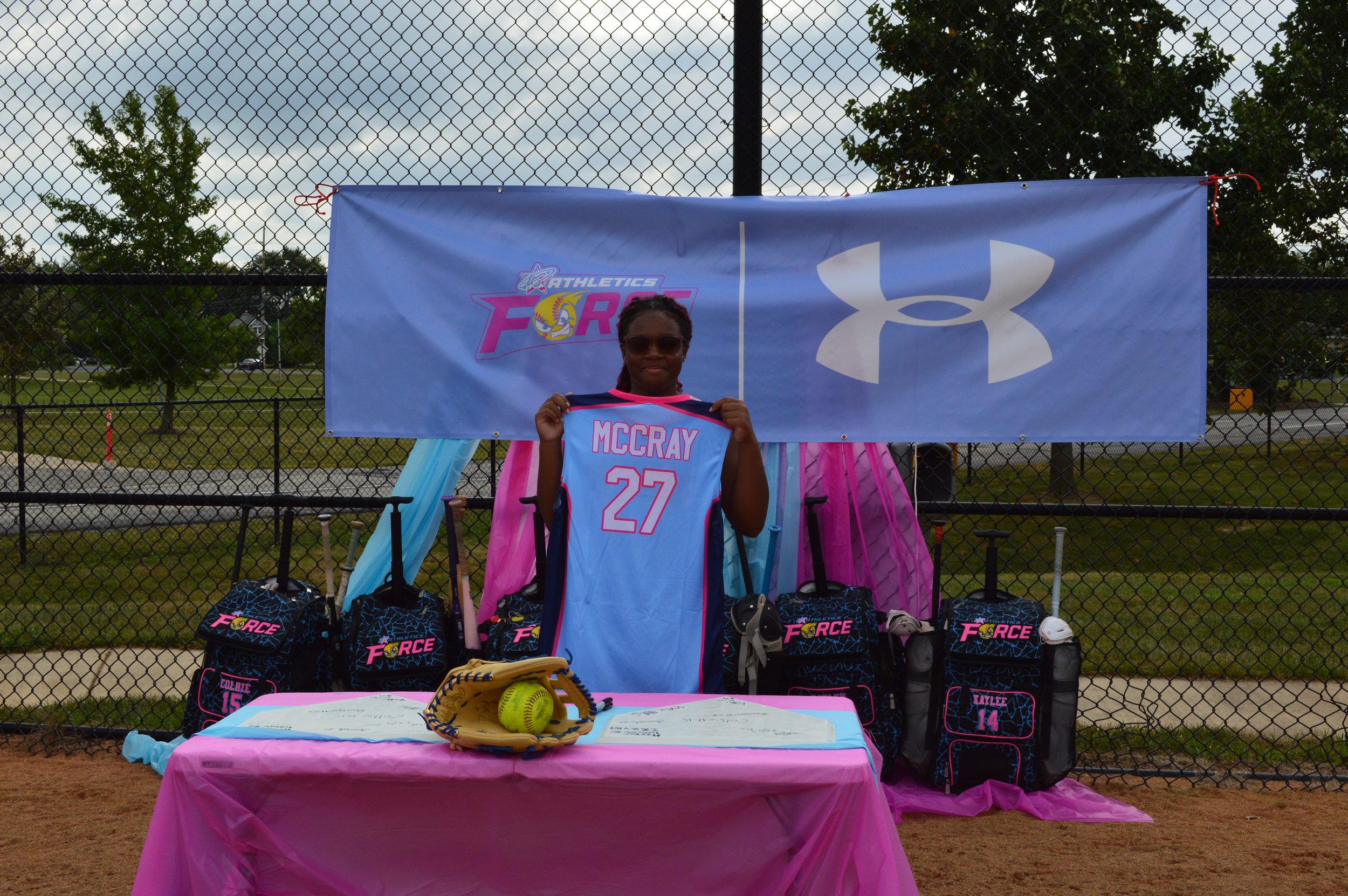 A woman holding a baseball jersey with the name 'McCray' and the number 27, standing in front of a blue banner for Athletics Force and Under Armour at a sports event. The table in front has sports gear, including a glove and a yellow softball.