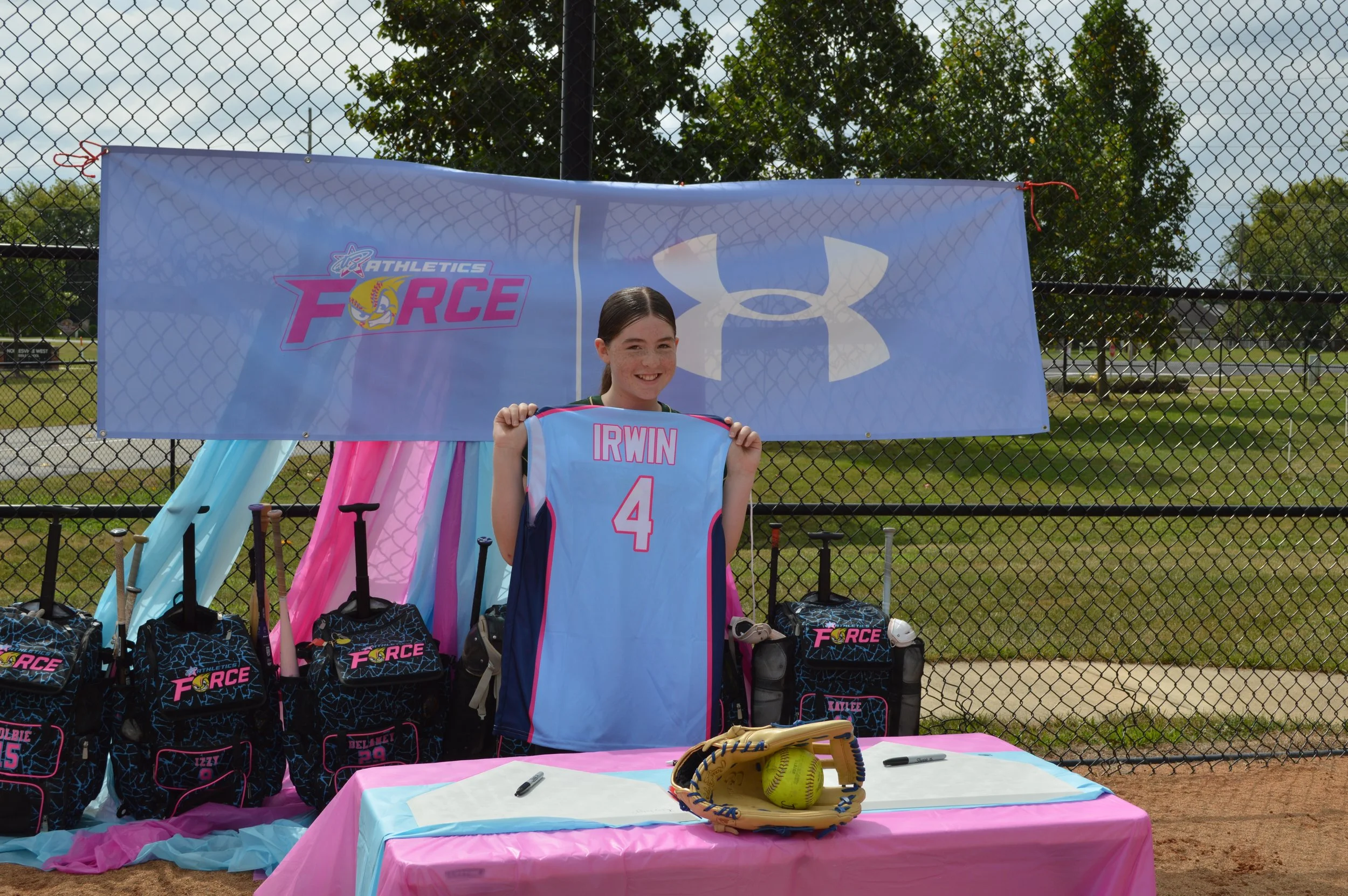 A young girl smiling at a youth softball event, holding up a softball jersey with her name 'IRWIN' and number 4. Behind her is a blue banner with logos for 'Athletics Force' and the Under Armour brand, a fence, trees, and a baseball field. There are sports bags, water bottles, and a softball glove on a table covered with pink and blue cloths.