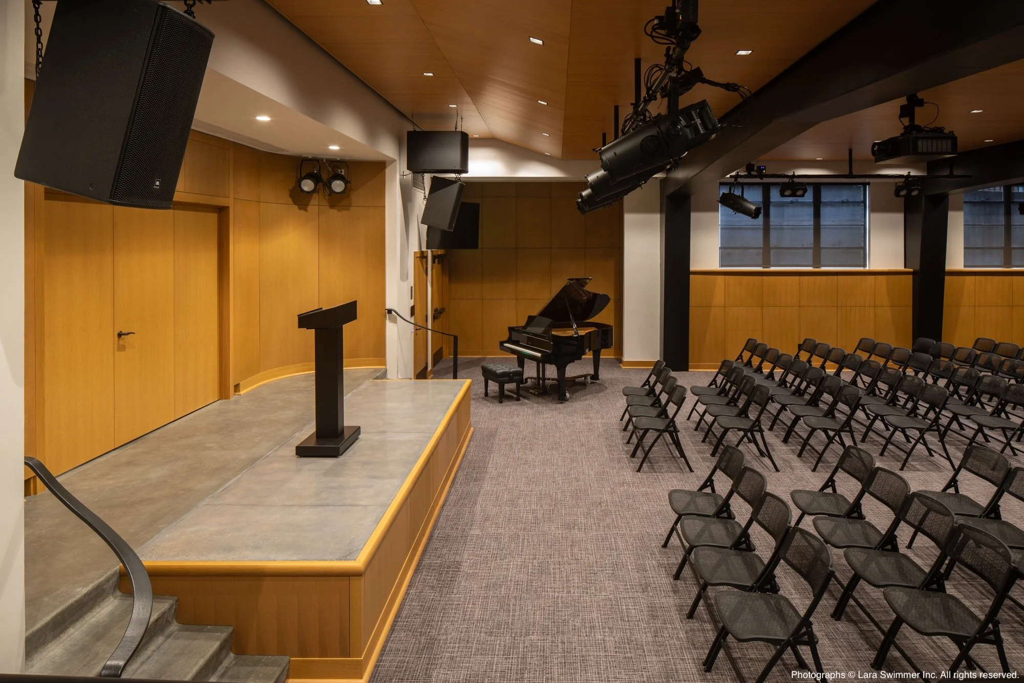 An empty auditorium or theater with a small stage, a microphone stand, a grand piano, chairs arranged in rows, and a podium. The space has wood-paneled walls, large windows, and overhead lighting.
