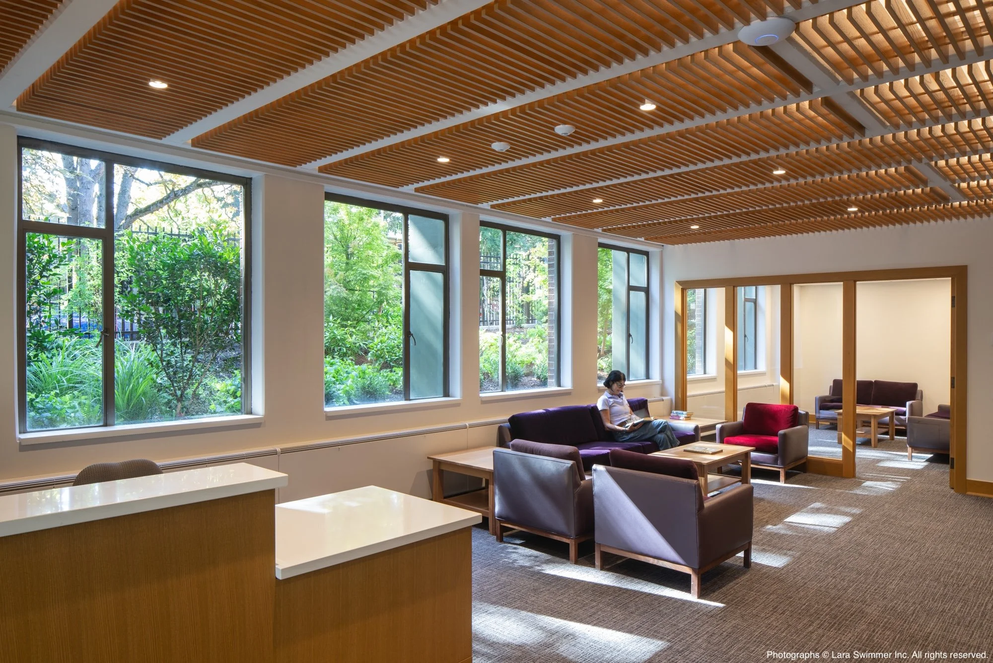 A modern indoor lounge area with large windows showing green trees outside. The ceiling has wooden slats with built-in lights. There are several chairs and sofas, with a woman sitting reading a book.