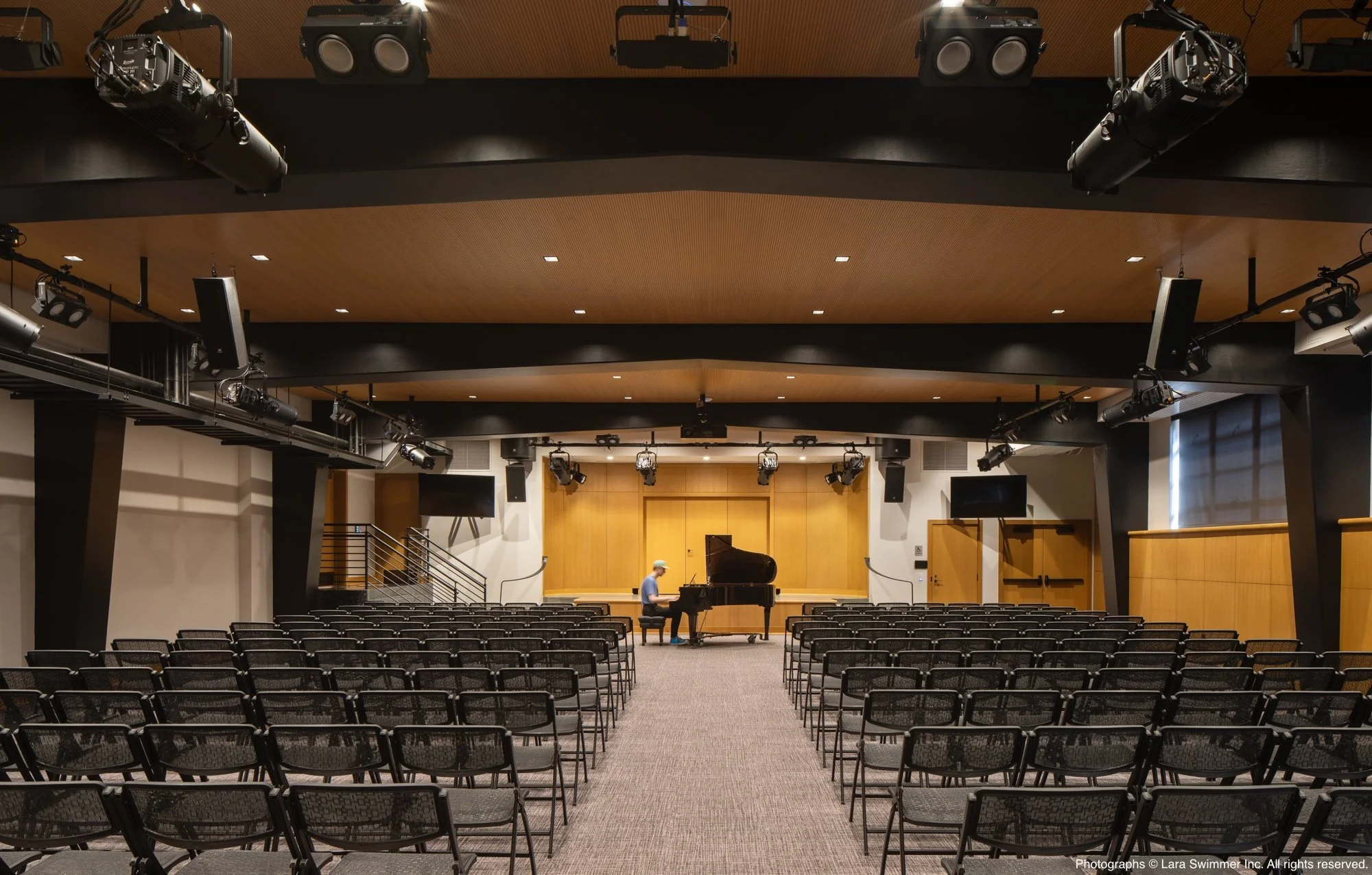 A concert hall or auditorium with rows of black seats, a stage with a grand piano, a person sitting at the piano, and a man standing nearby. The hall has a wooden ceiling with ceiling lights and mounted speakers.