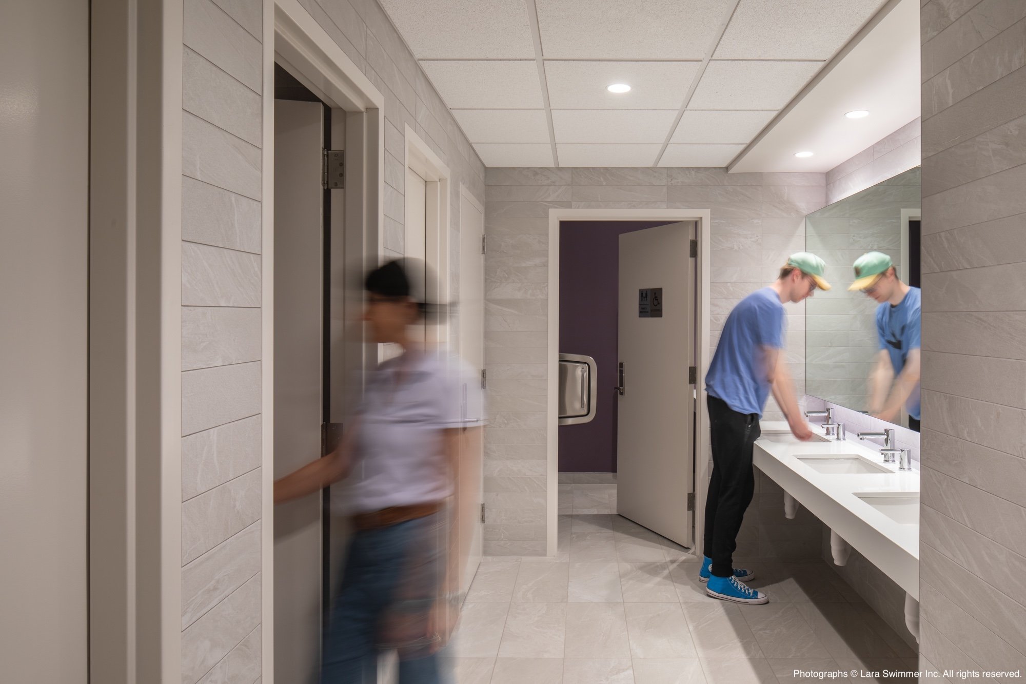 Two young men wearing blue shirts and green caps washing their hands at a white sink in a public restroom with beige tiled walls and a large mirror.