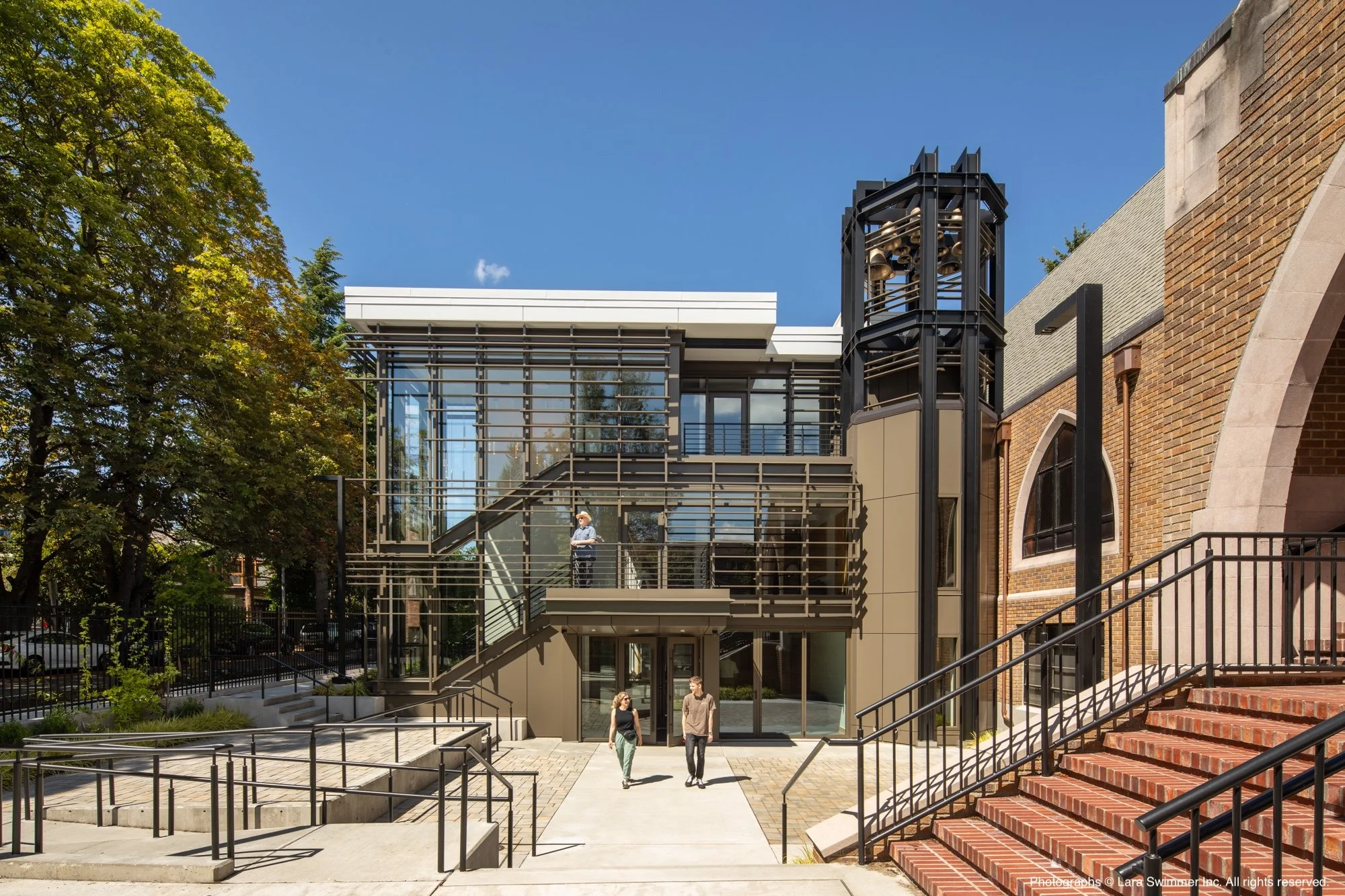 People walking outside a modern building with glass and metal features, a bell tower, and brick walls under a clear blue sky.