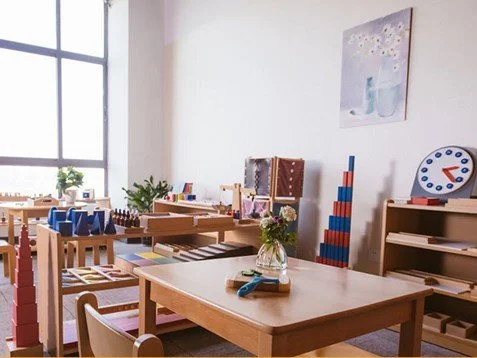 Classroom with wooden tables, chairs, shelves, educational toys, and a large window with plants outside.