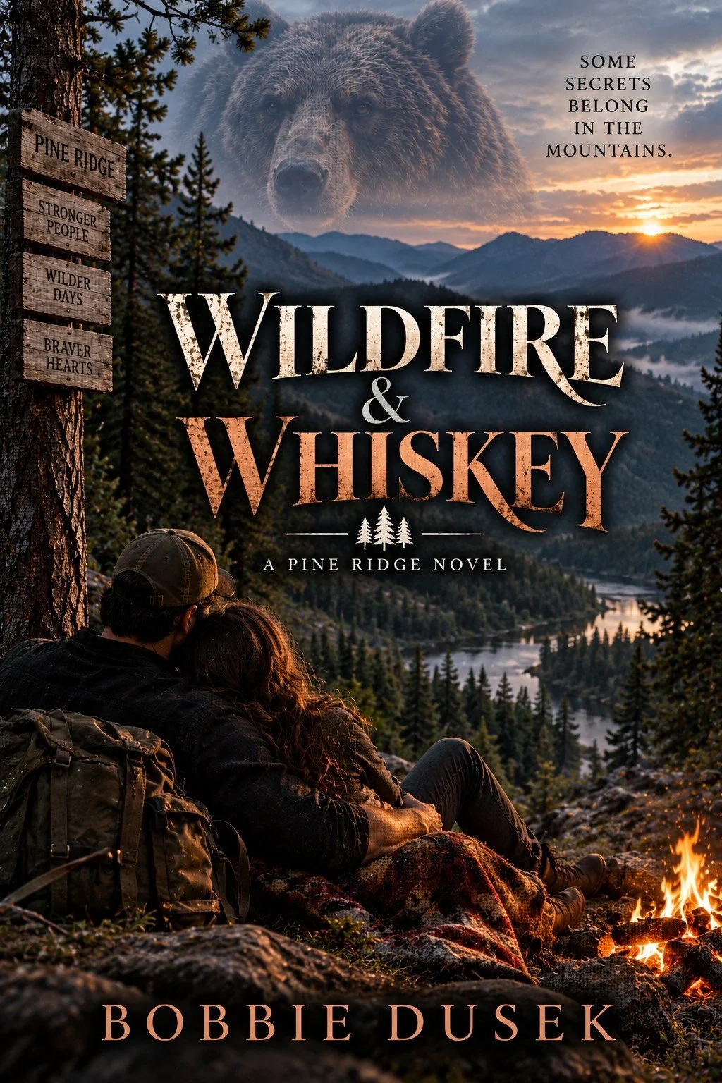 Book cover titled "Wildfire & Whiskey" by Bobbie Dusek. Features a couple sitting by a campfire in a forest with pine trees and a river, overlooking mountains and a sunset. A bear's face appears in the sky along with the quote "Some secrets belong in the mountains." There is a wooden sign with the words "Pine Ridge," "Stronger People," "Wilder Days," and "Braver Hearts."