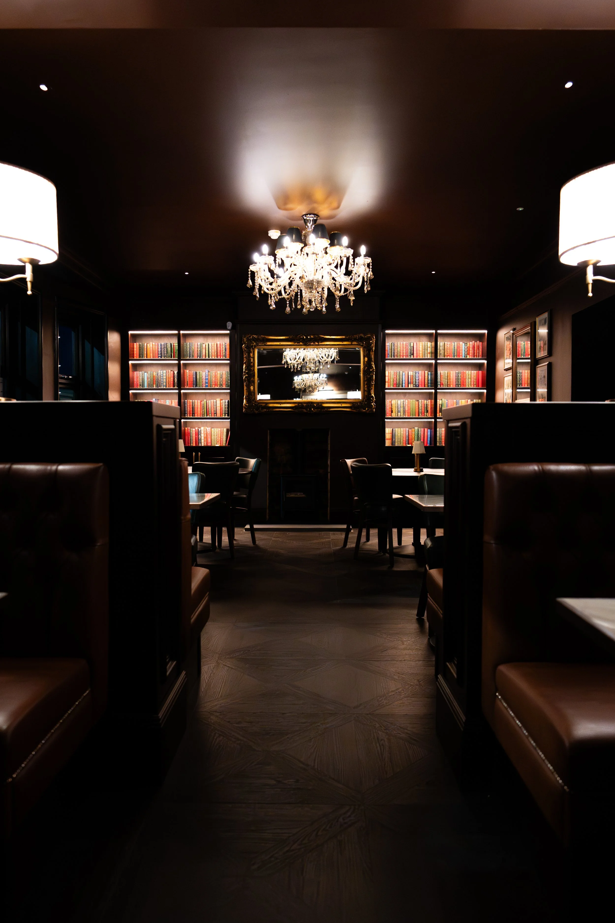 A luxurious library with dark walls, illuminated bookshelves filled with colorful books, a large ornate mirror, chandelier, and elegant seating areas.