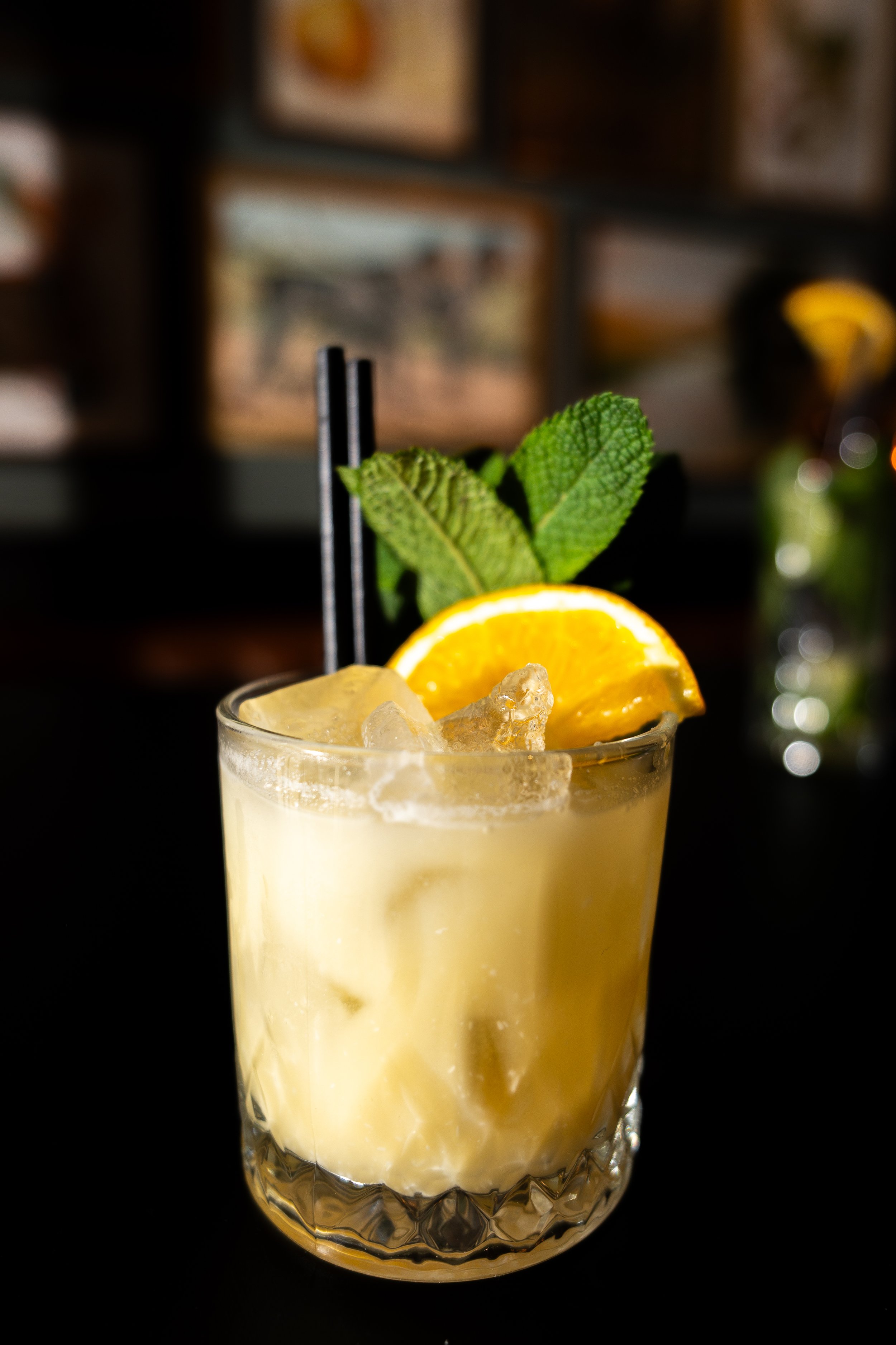 A close-up of a yellow cocktail in a glass with ice, garnished with a slice of orange, mint leaves, and black stir sticks, against a blurred dark background.