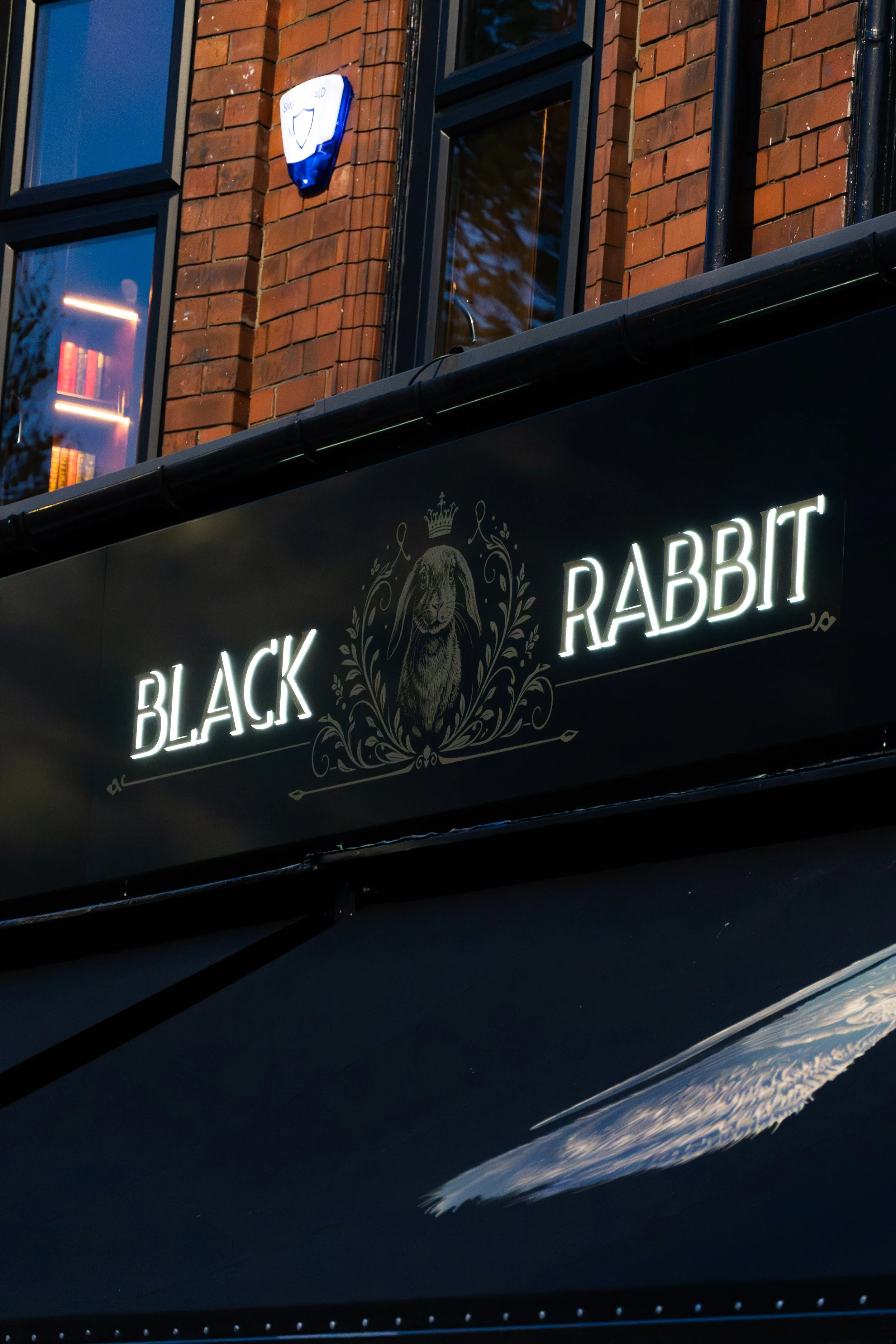 Nighttime street scene with illuminated black sign reading 'Black Rabbit' and a white rabbit logo, brick building with window, and blue and white emergency shield on the wall.