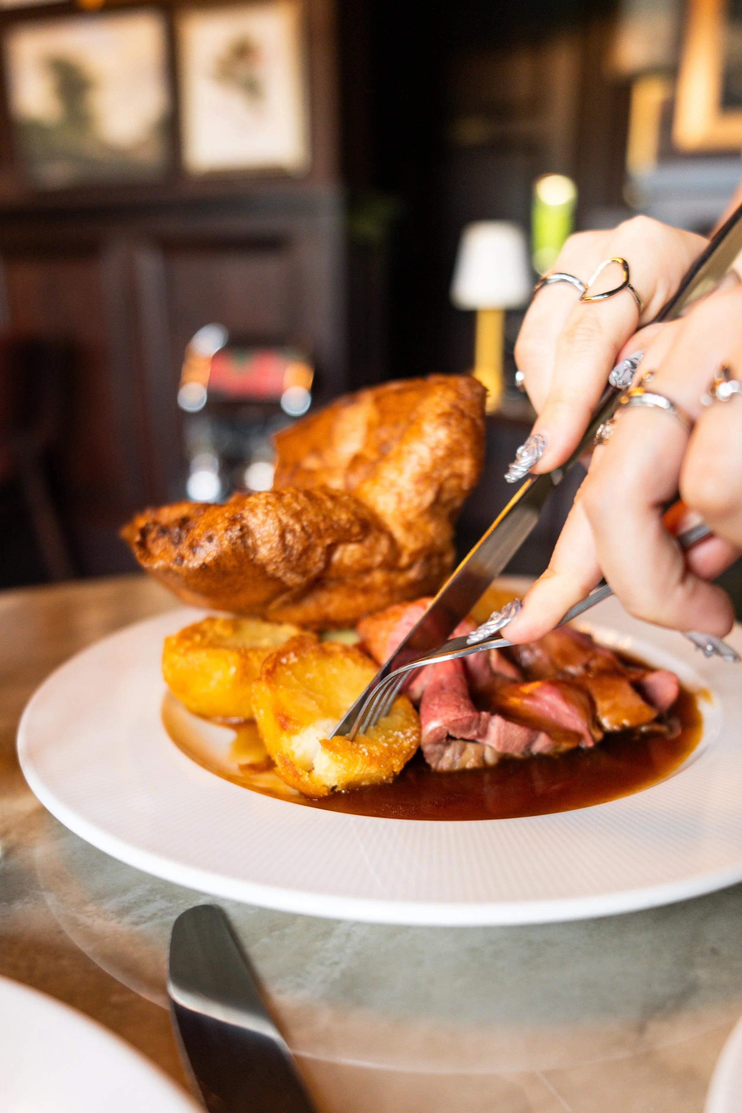 A plate of roasted meat, potatoes, and vegetables, with a large piece of fried chicken being cut by a person with rings on their fingers in a cozy restaurant setting.