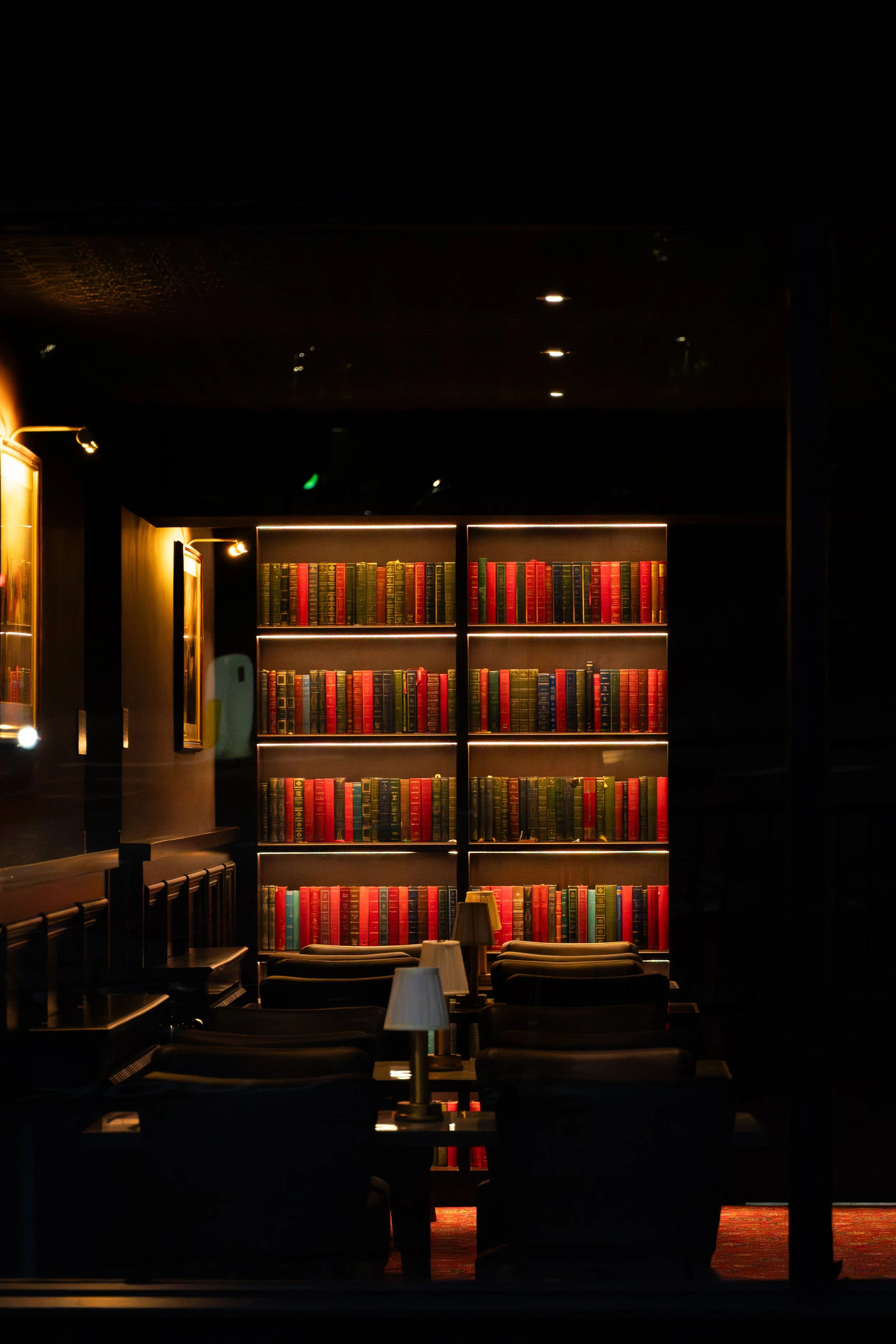A library interior with dark walls, illuminated by warm lighting. Focused on a large bookshelf filled with multicolored books, and seating arrangements with chairs and tables in the foreground.