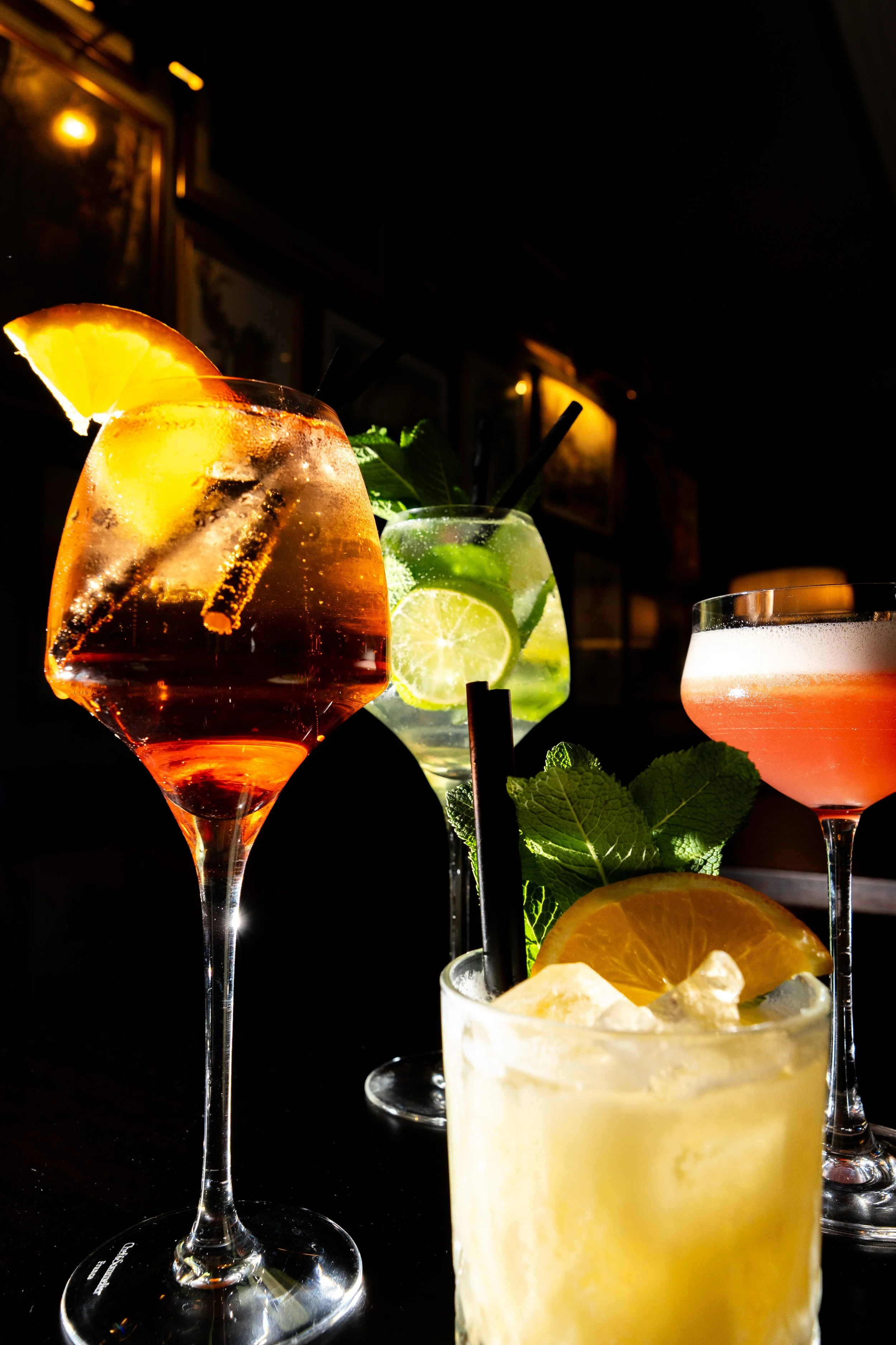 Four colorful cocktails on a dark bar counter with a black background. The drinks have garnishes of citrus slices, mint leaves, and ice, with straws in each glass.