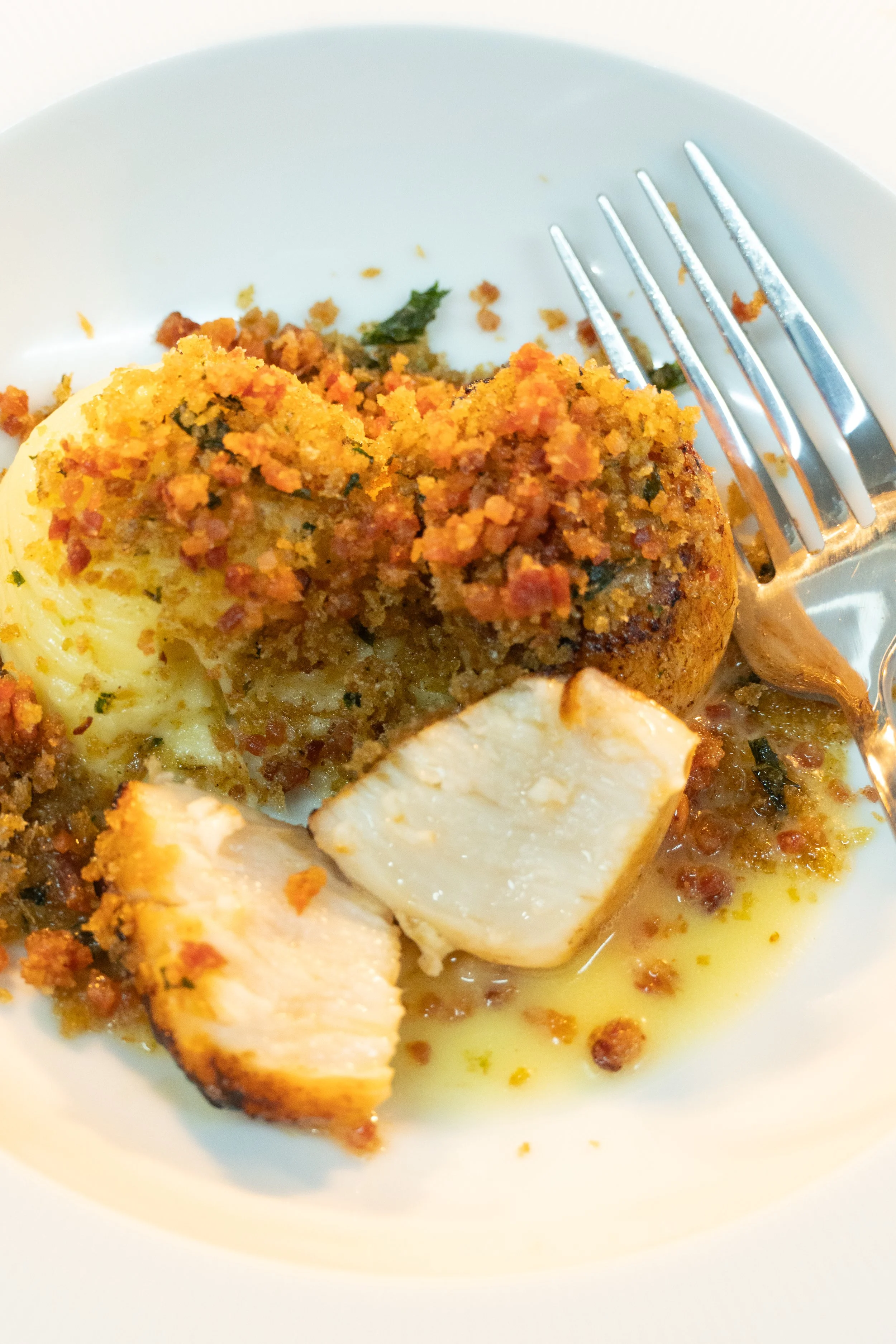 Close-up of a plate with a piece of cooked fish topped with seasoned bread crumbs, garnished with herbs, alongside mashed potatoes with herbs, and a fork resting on the side of the plate