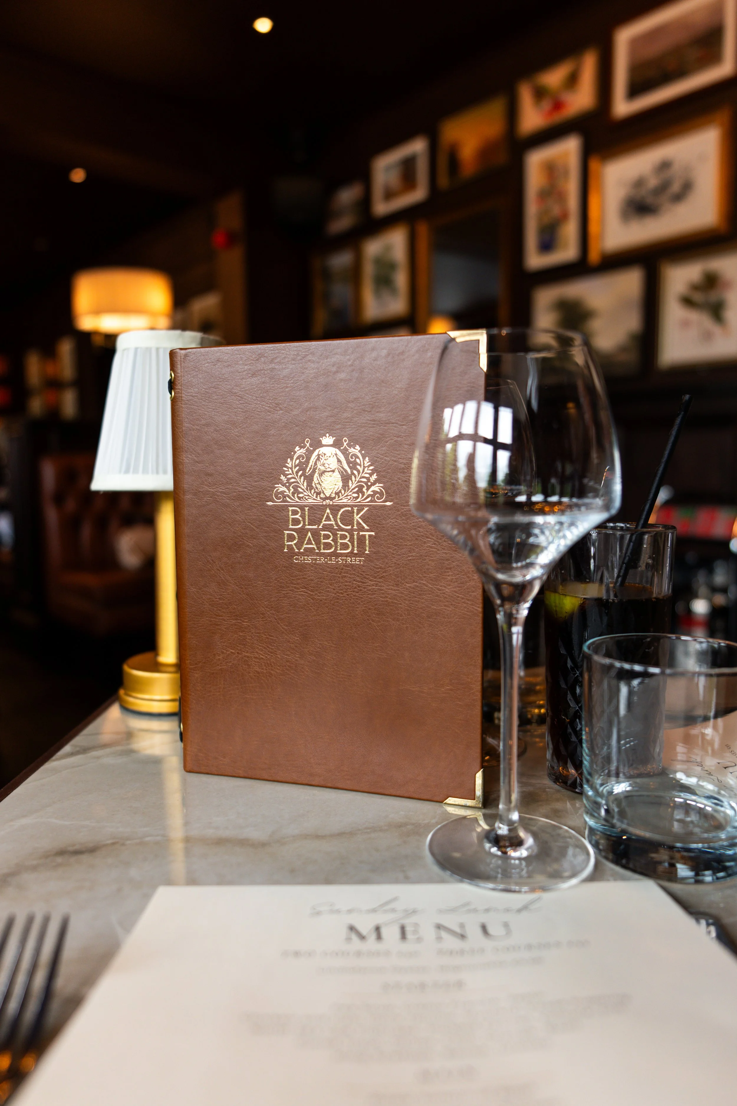 A restaurant table setting with a menu from Black Rabbit Chester-Le-Street, a wine glass, a glass with a straw, and a lit lamp, with framed artwork on a dark wall in the background.