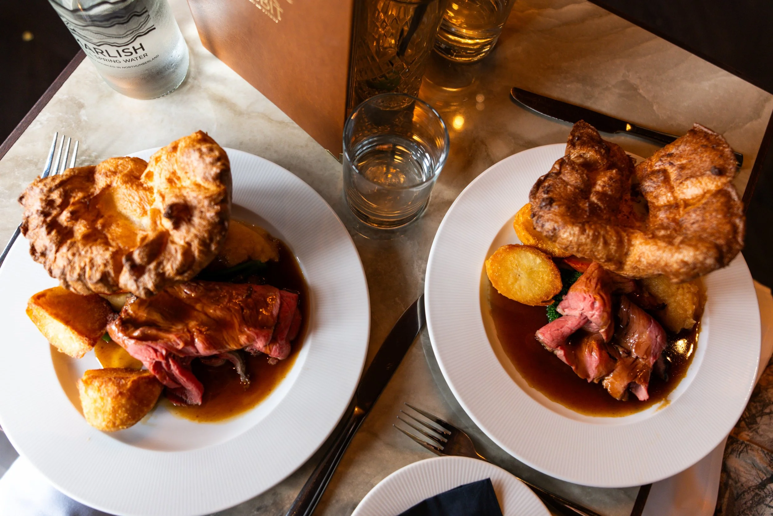 Two plates of roast beef with Yorkshire pudding, roast potatoes, and vegetables on a table set for a meal, with cutlery, a glass of water, and a water bottle.