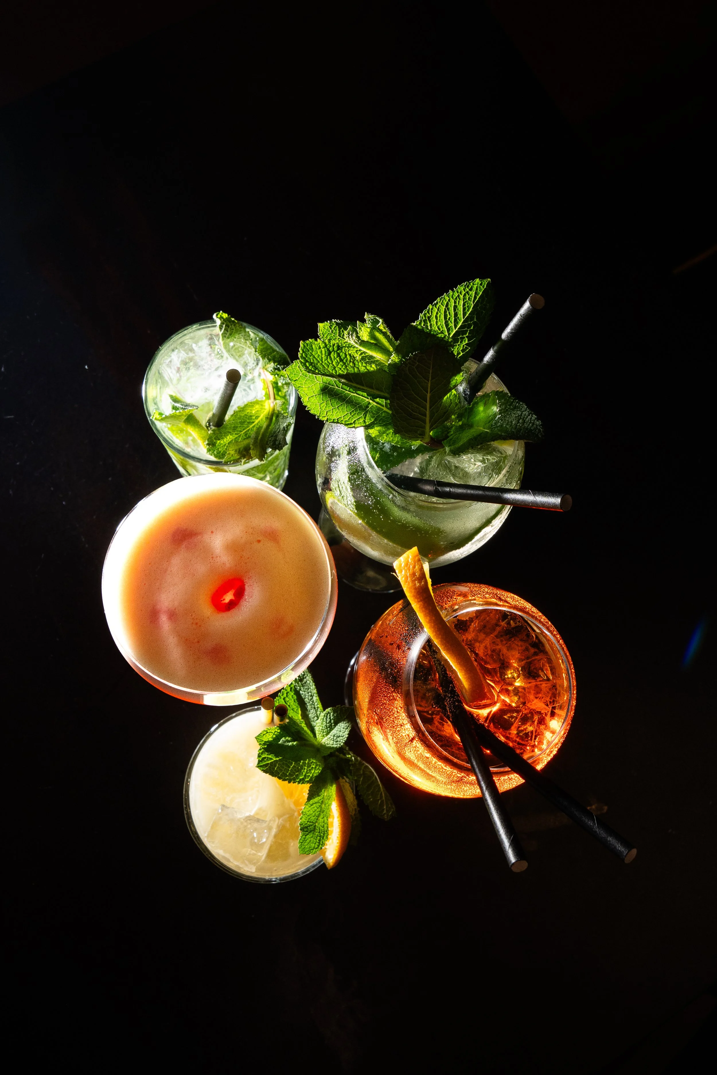 Top-down view of four colorful cocktails with mint leaves and lemon slices, four black straws, against a dark background.