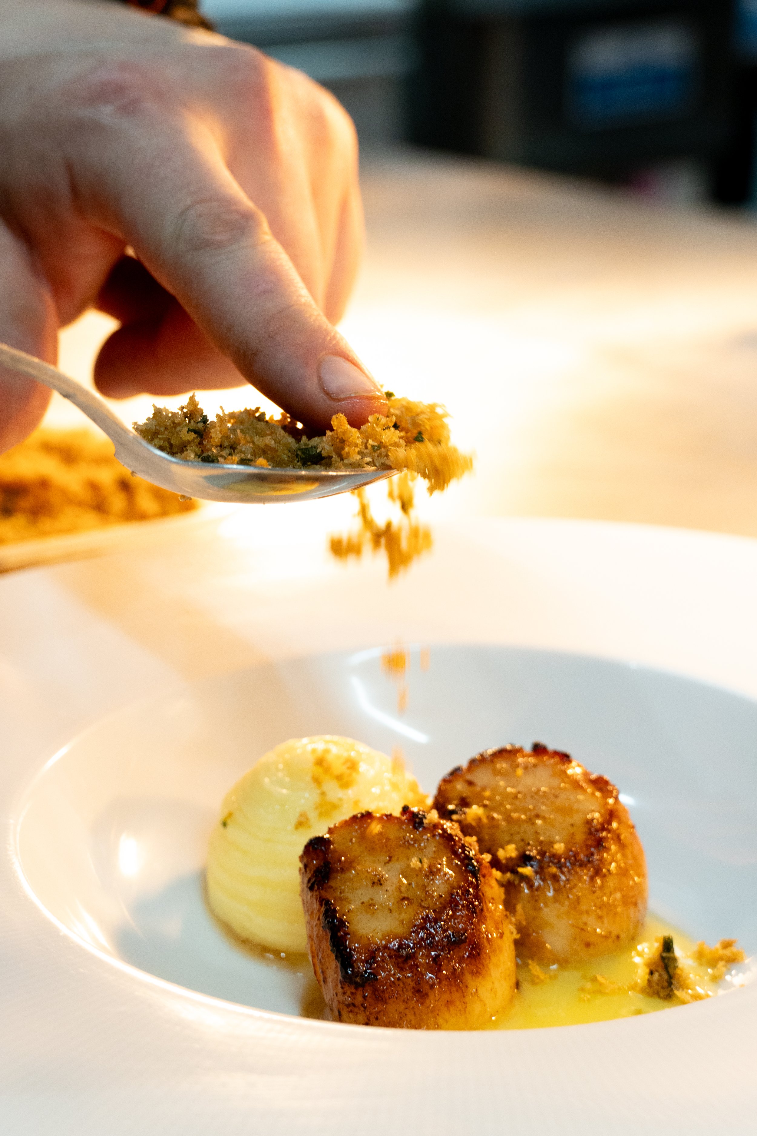 Close-up of a hand sprinkling herb atop seared scallops and mashed potatoes on a white plate.